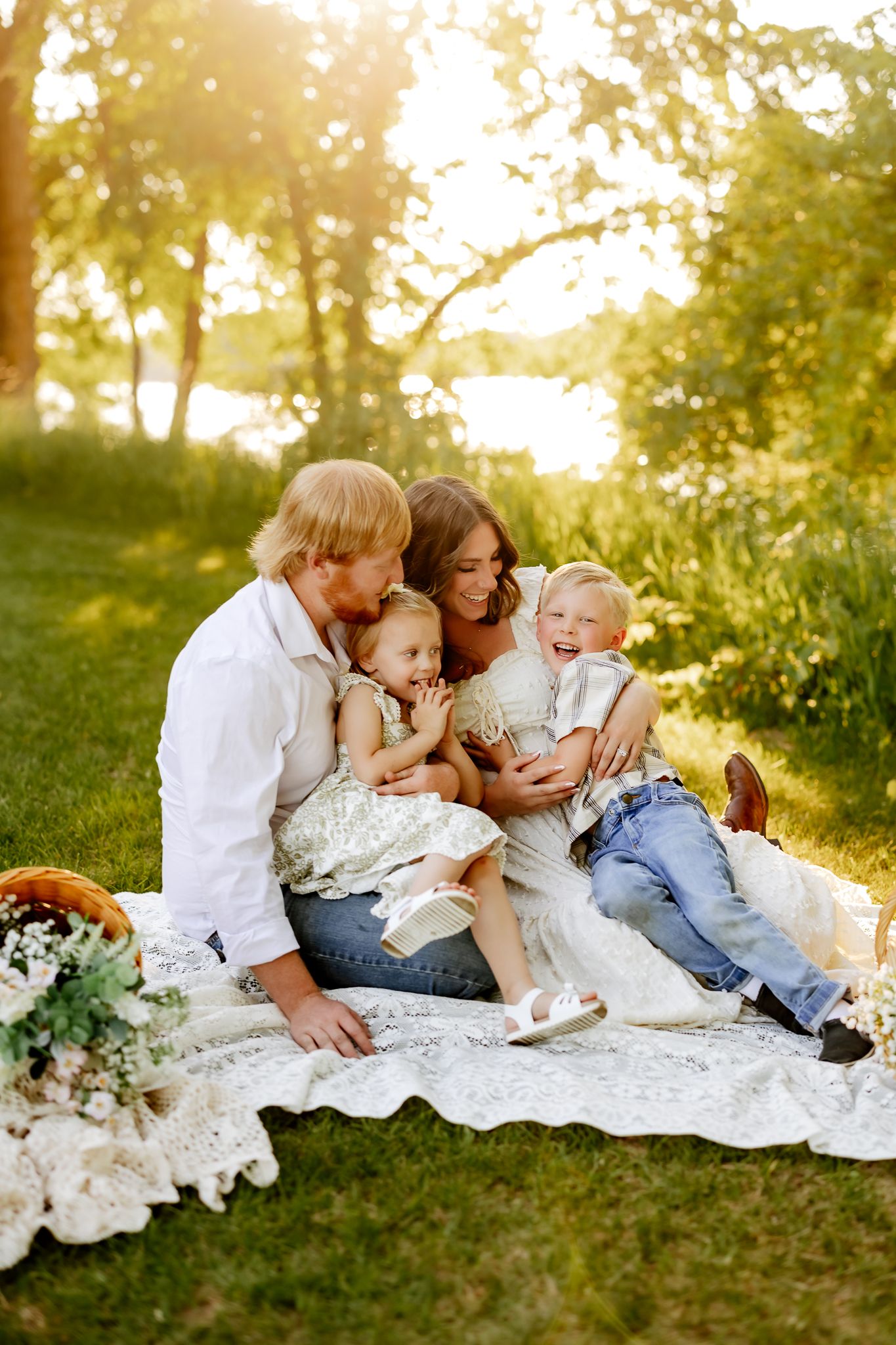 Happy family sitting on lace blanket in sunny park, parents laughing with two young children during outdoor picnic.