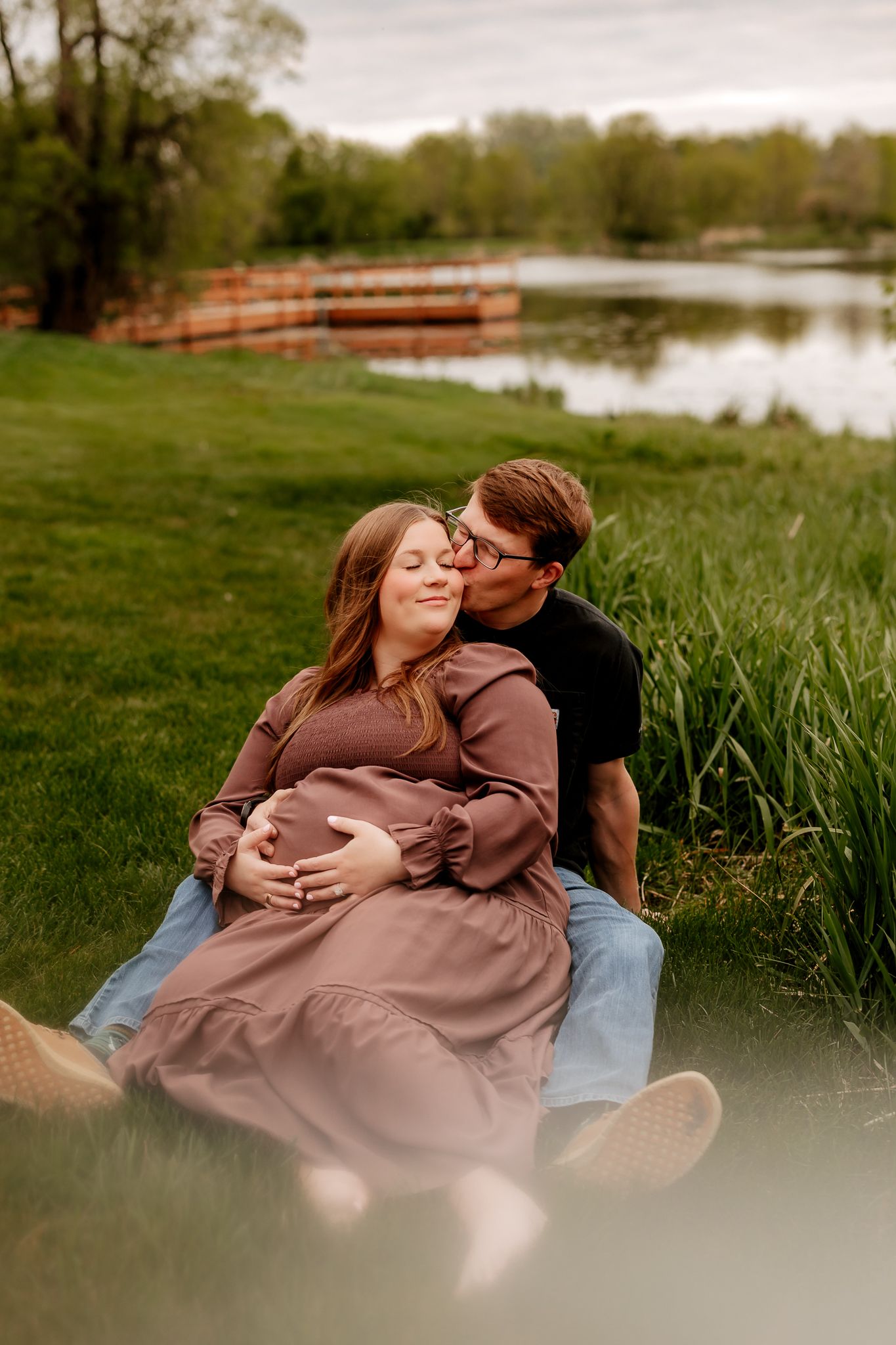 A couple sits on grass by a pond, the man kissing a pregnant woman on the cheek at dusk.