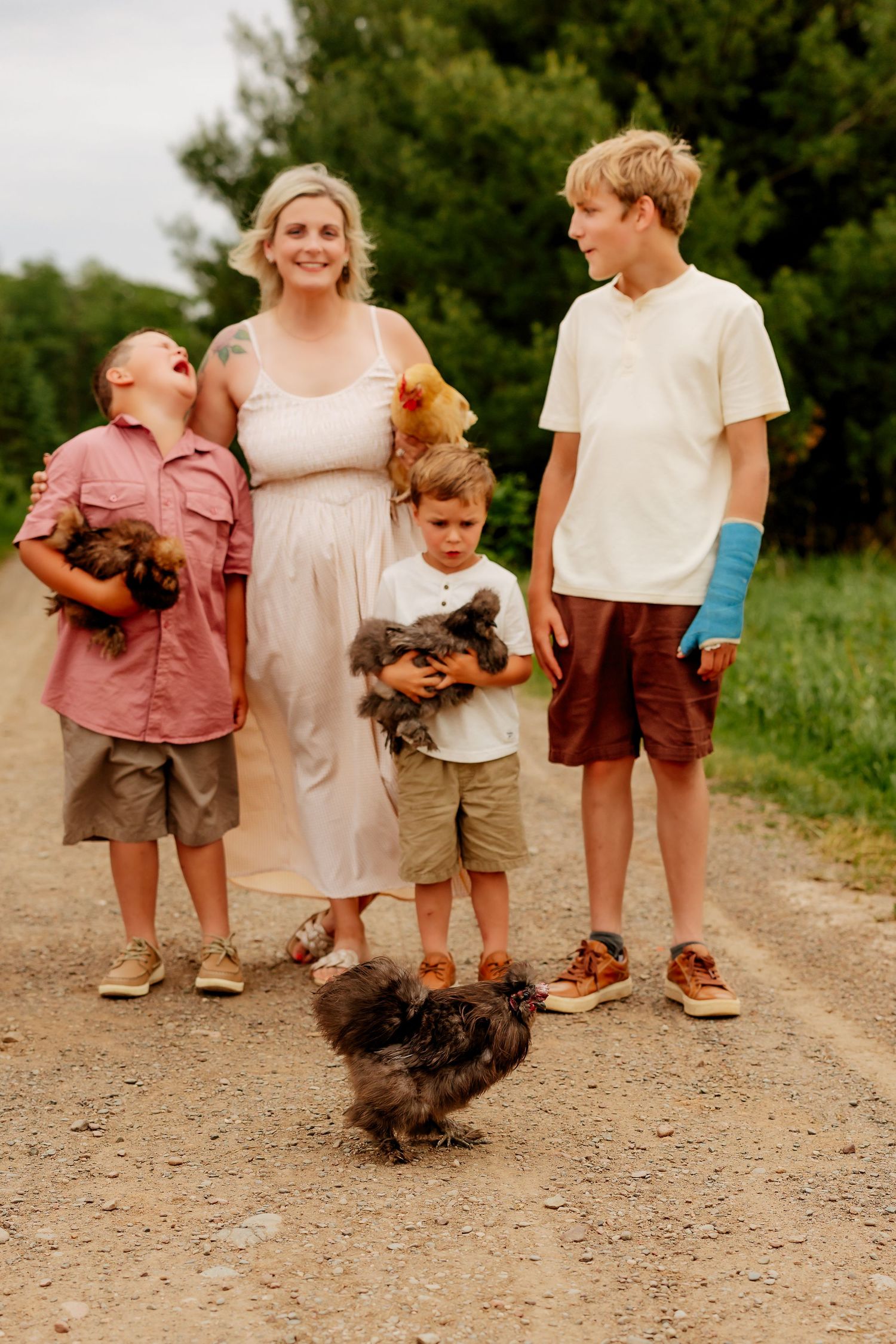 Smiling mother with three boys holding chickens on a rural dirt road surrounded by greenery.