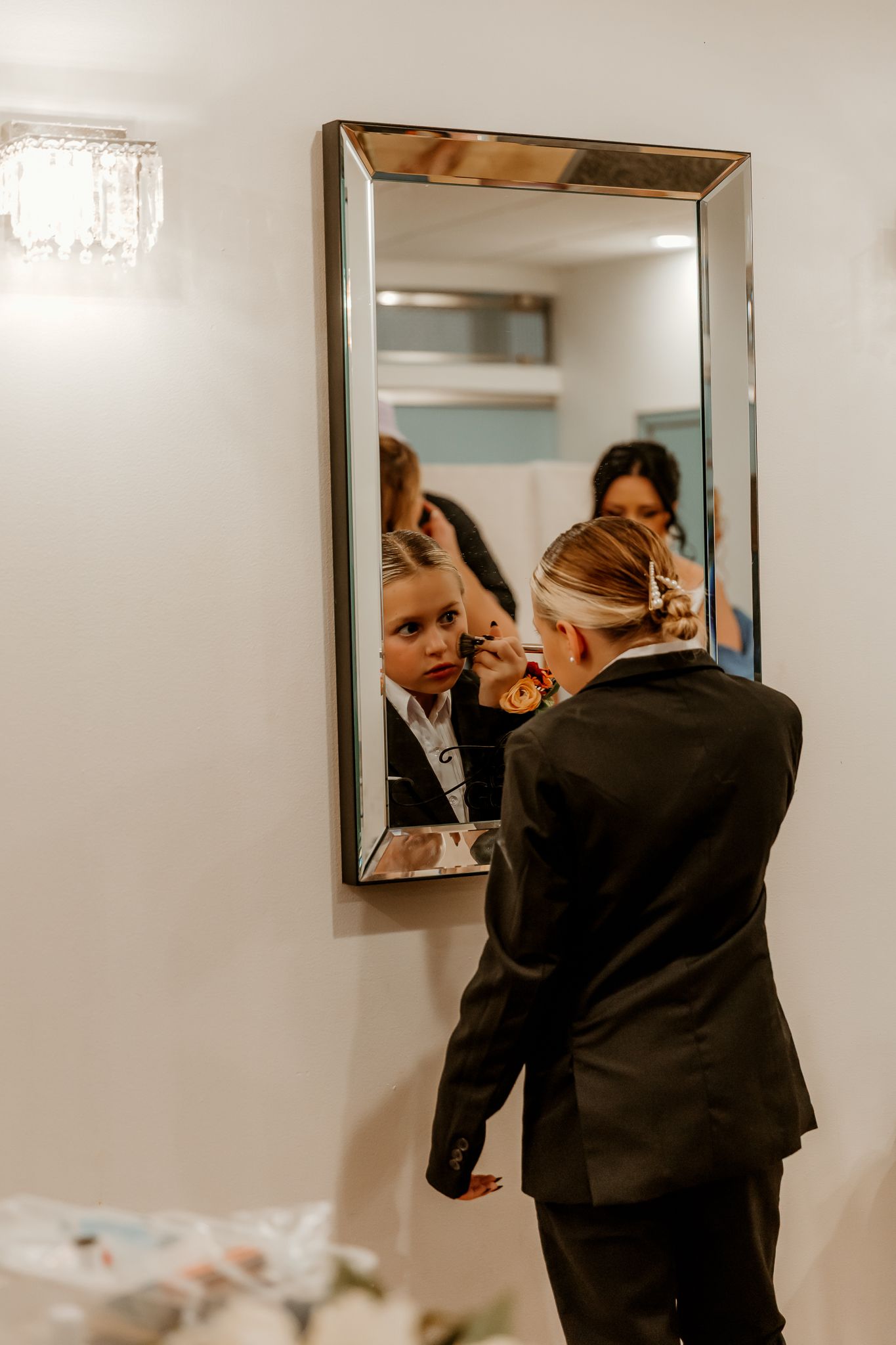 Woman in a dark blazer with hair in an updo applies makeup while looking into a framed mirror that reflects other women getting ready.