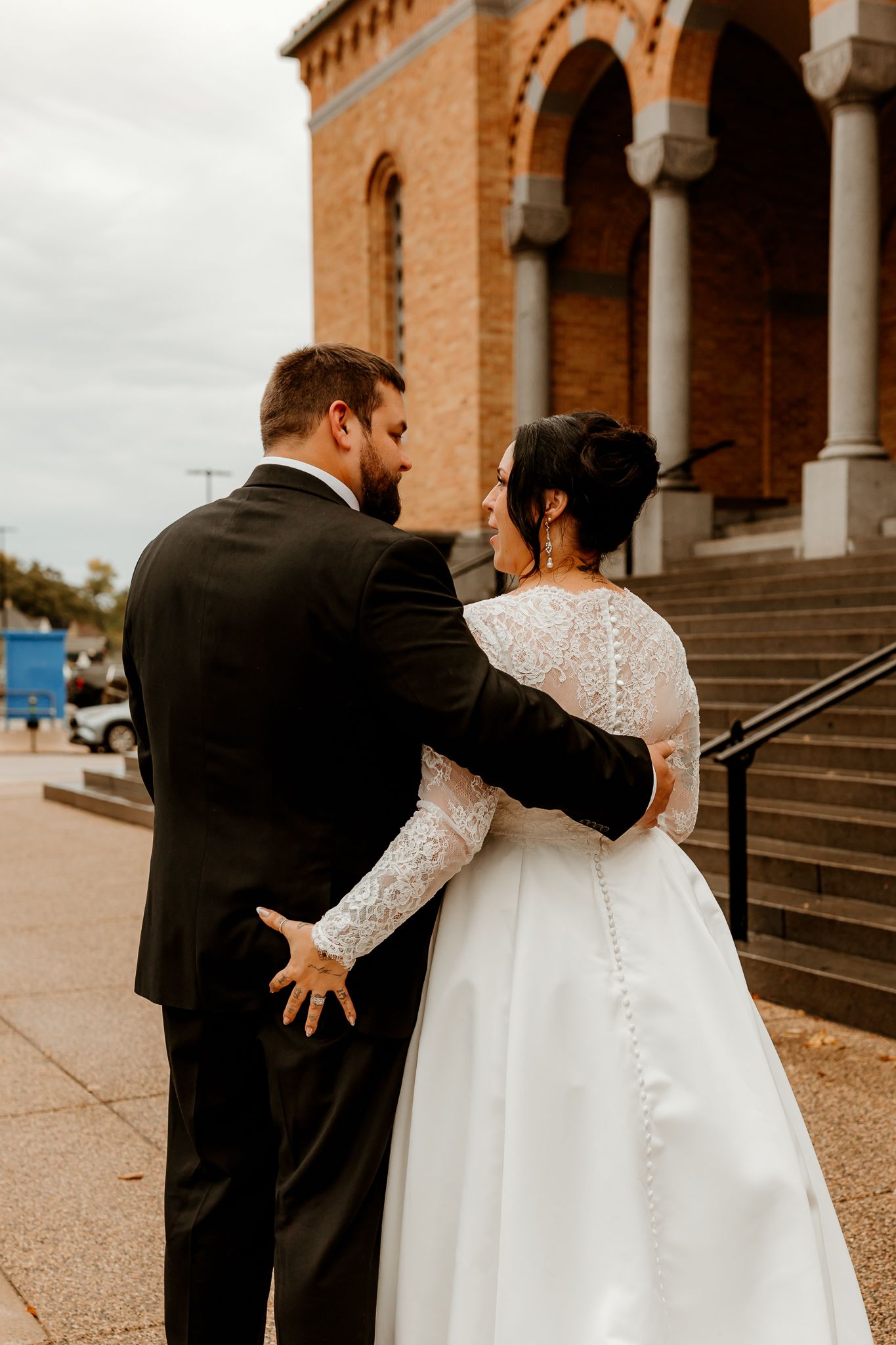 Bride in a lace-sleeved white wedding gown and groom in a dark suit embrace outside a brick church with arched windows and stone steps.