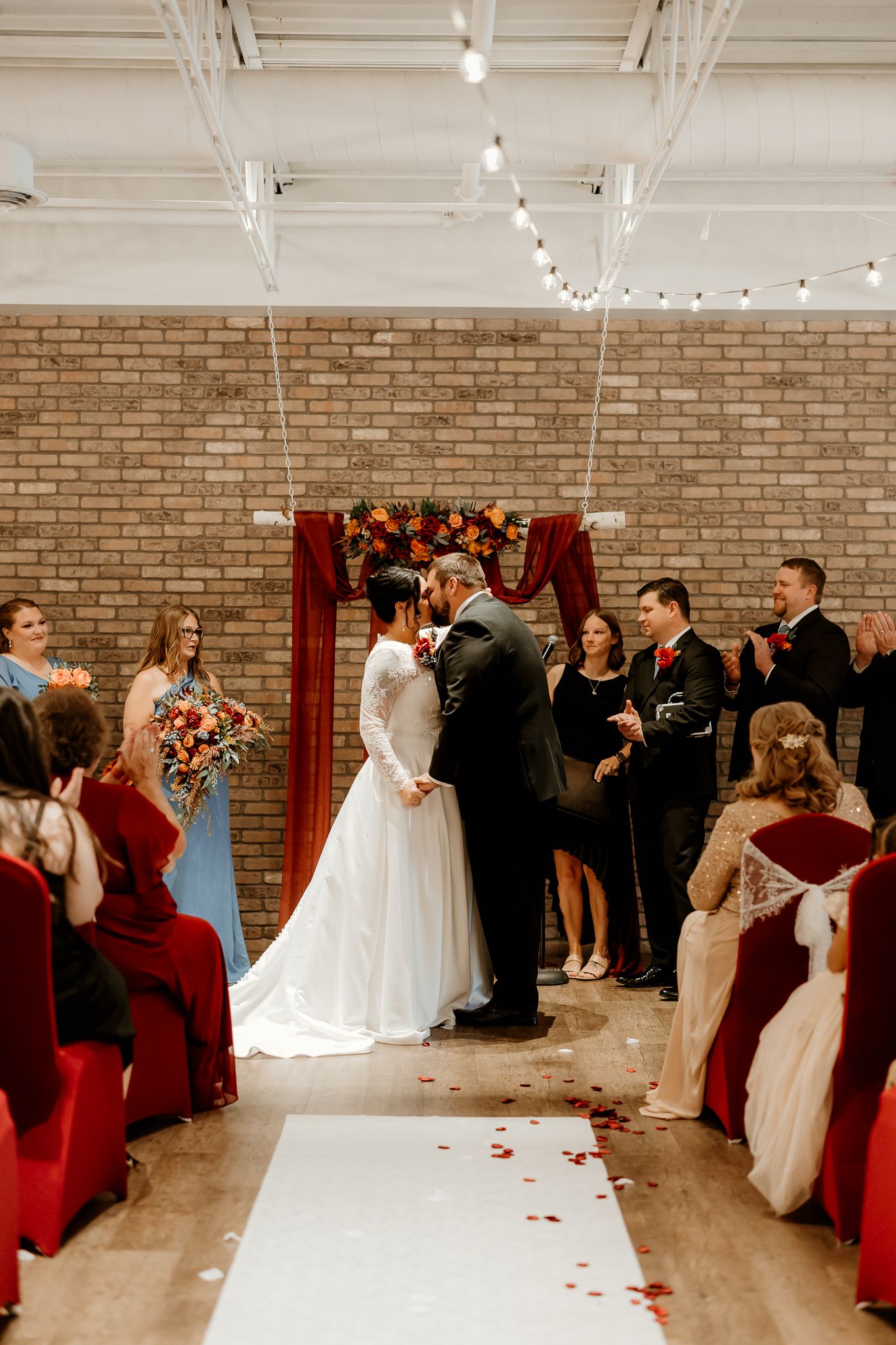 Bride in a white lace gown kisses the groom at an indoor brick-walled wedding ceremony as guests and groomsmen look on.