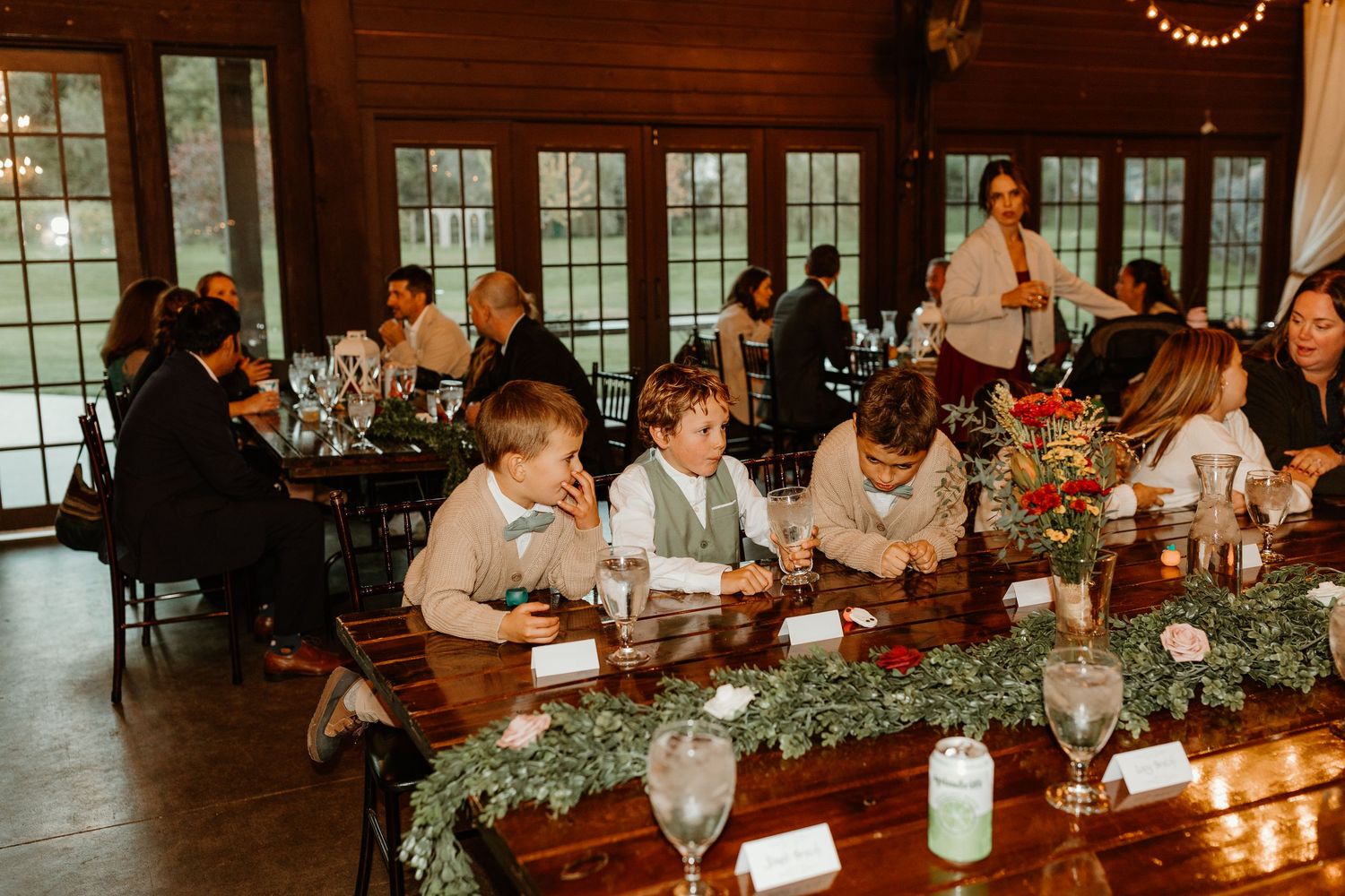 Three boys in dress shirts and vests sit at a long wedding table with greenery garland, candles, and glasses as guests mingle nearby.