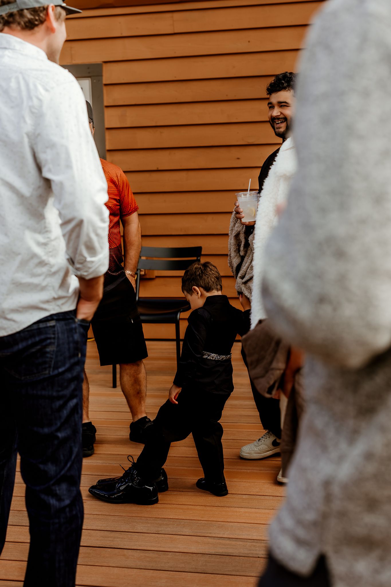 Young boy in black clothes and dress shoes dances playfully on a wooden deck while smiling guests stand around him at a casual gathering.