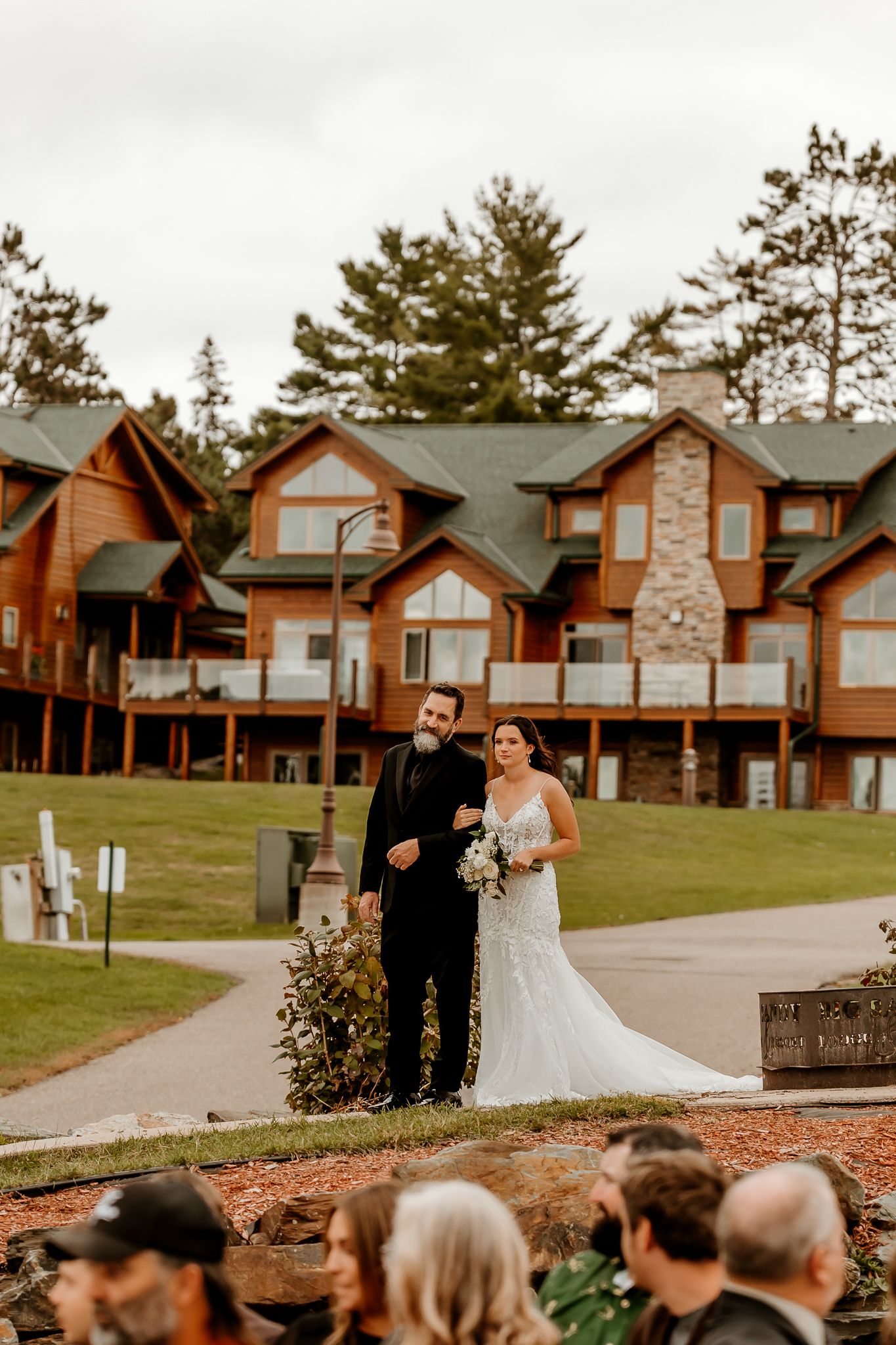 Bride in a lace wedding gown holding a bouquet walks arm in arm with a man in a dark suit before guests at a lakeside wooden lodge.