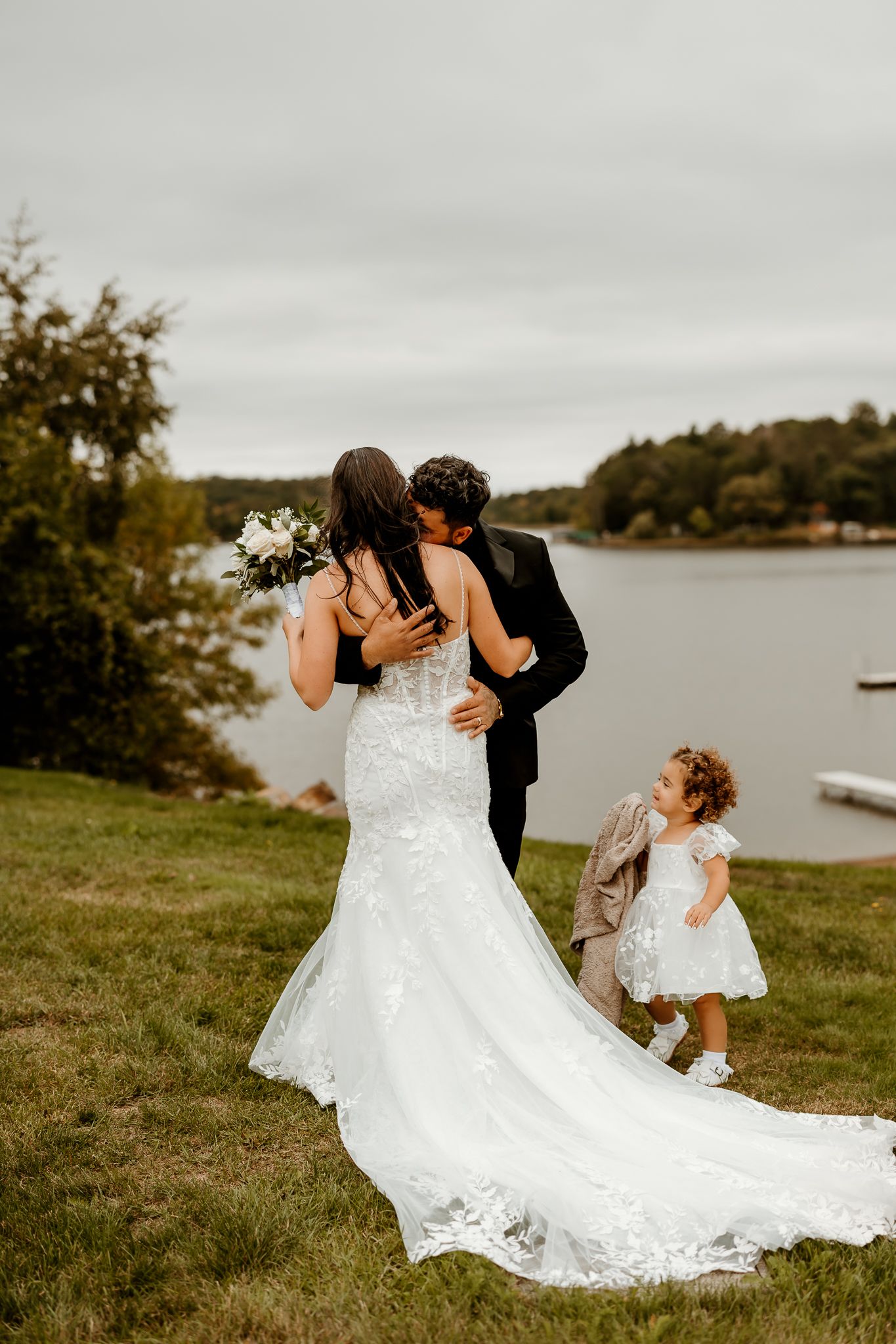 Bride in a lace wedding gown embraces her groom by a lake as a young flower girl in a white dress watches nearby.