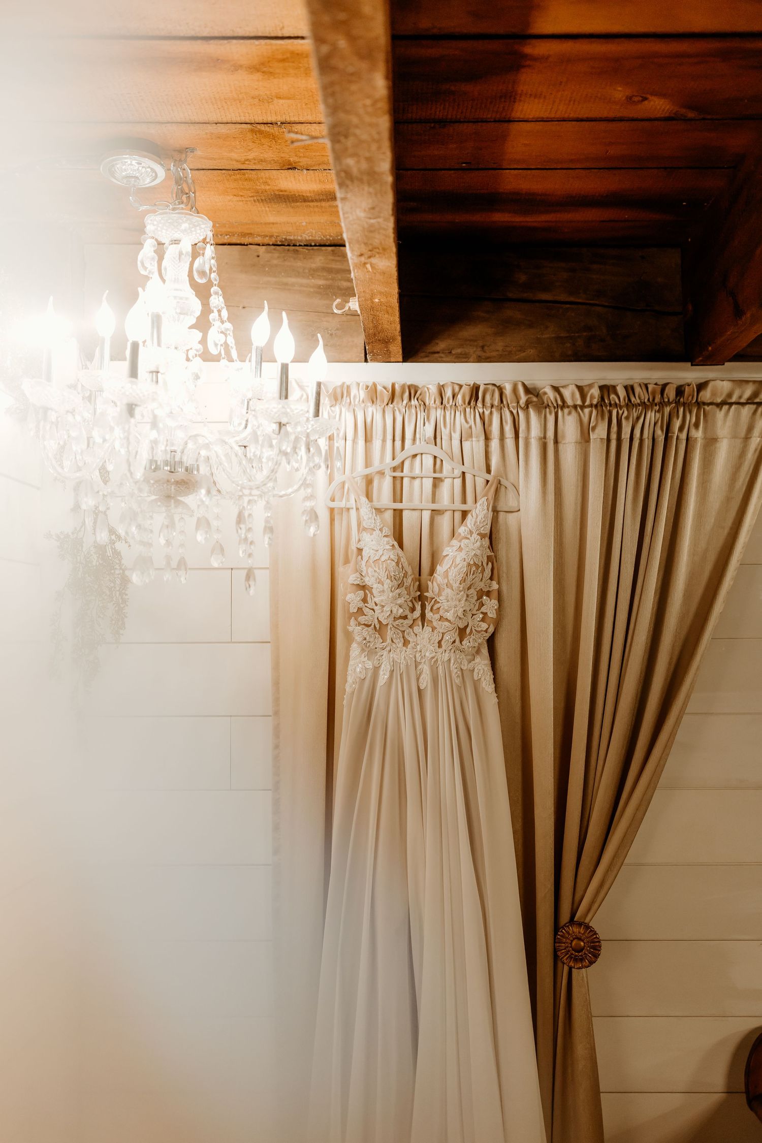 Lace-bodice wedding dress with spaghetti straps hanging beside a crystal chandelier in a rustic room with wood ceiling and shiplap walls.