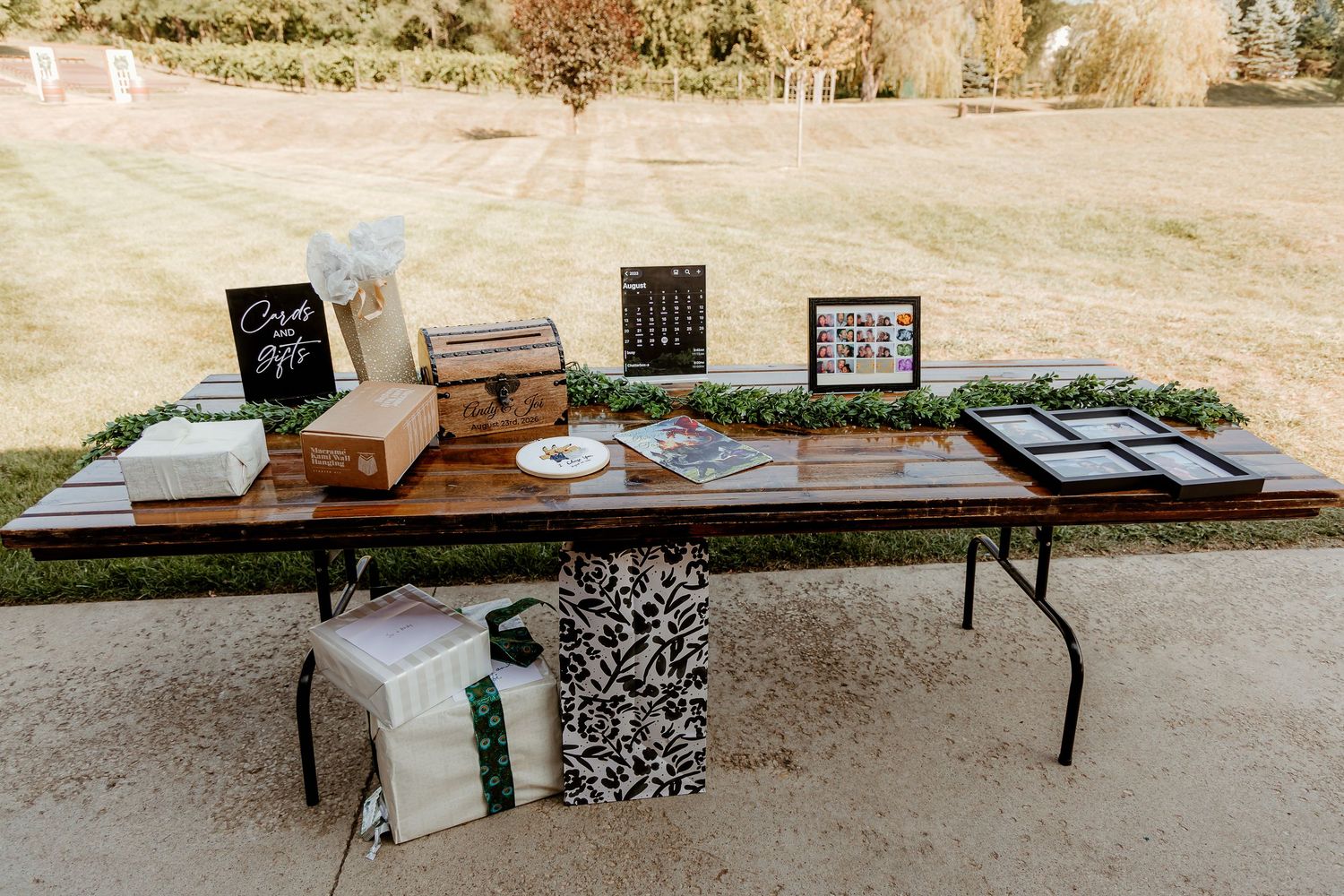Outdoor wedding gift table with suitcase card box, framed photos, greenery garland, and wrapped presents on a rustic wood folding table.