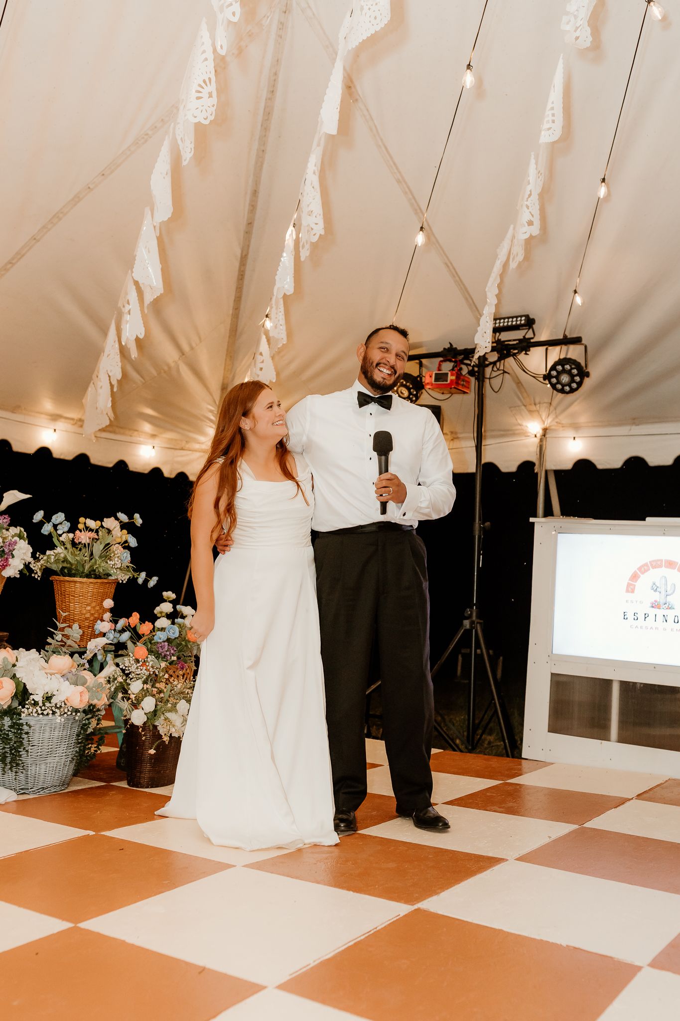 A bride and groom stand together on a checkered dance floor in a decorated tent during their wedding reception.