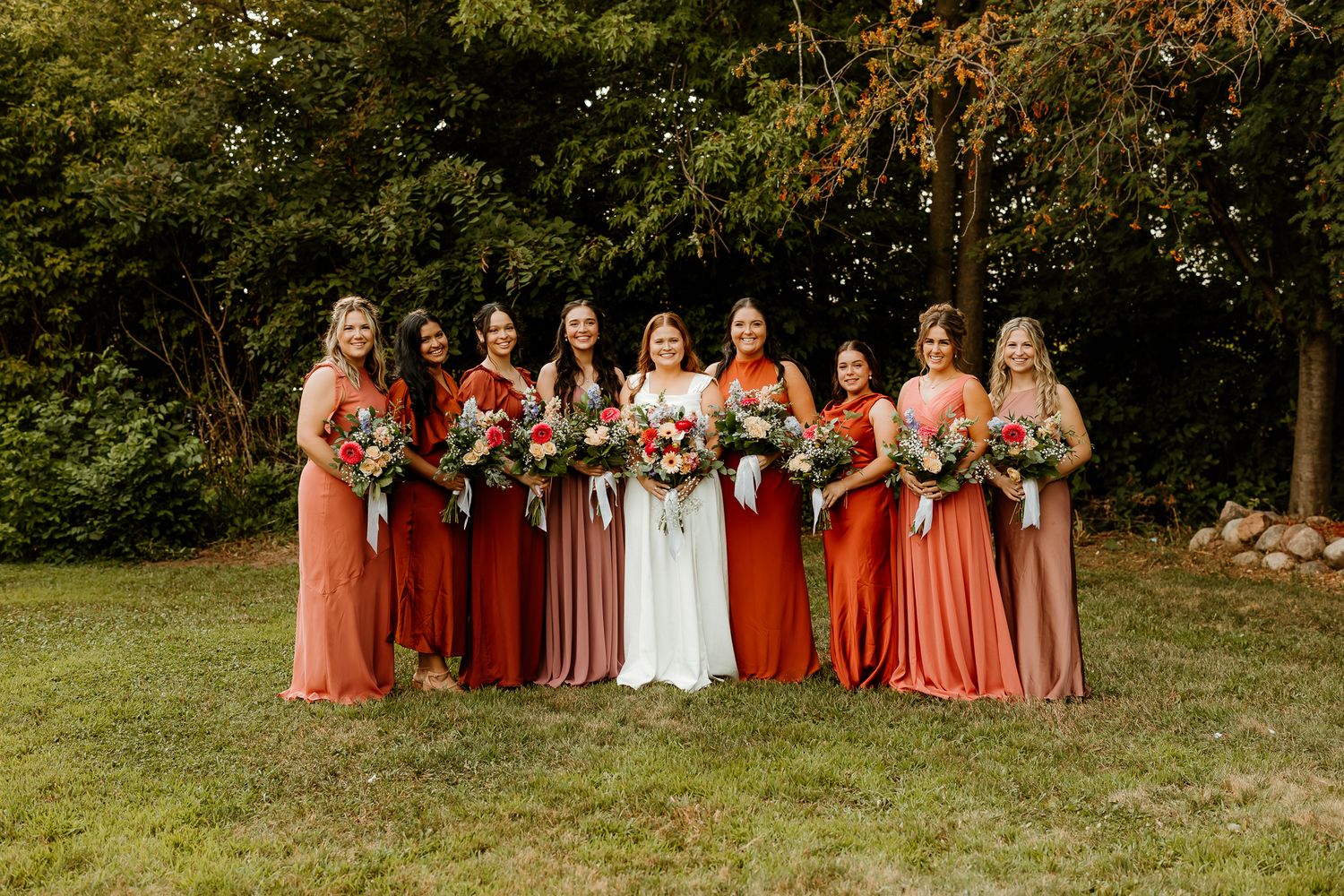 Bride in white gown poses with eight bridesmaids in mismatched rust and coral dresses holding bouquets outdoors.