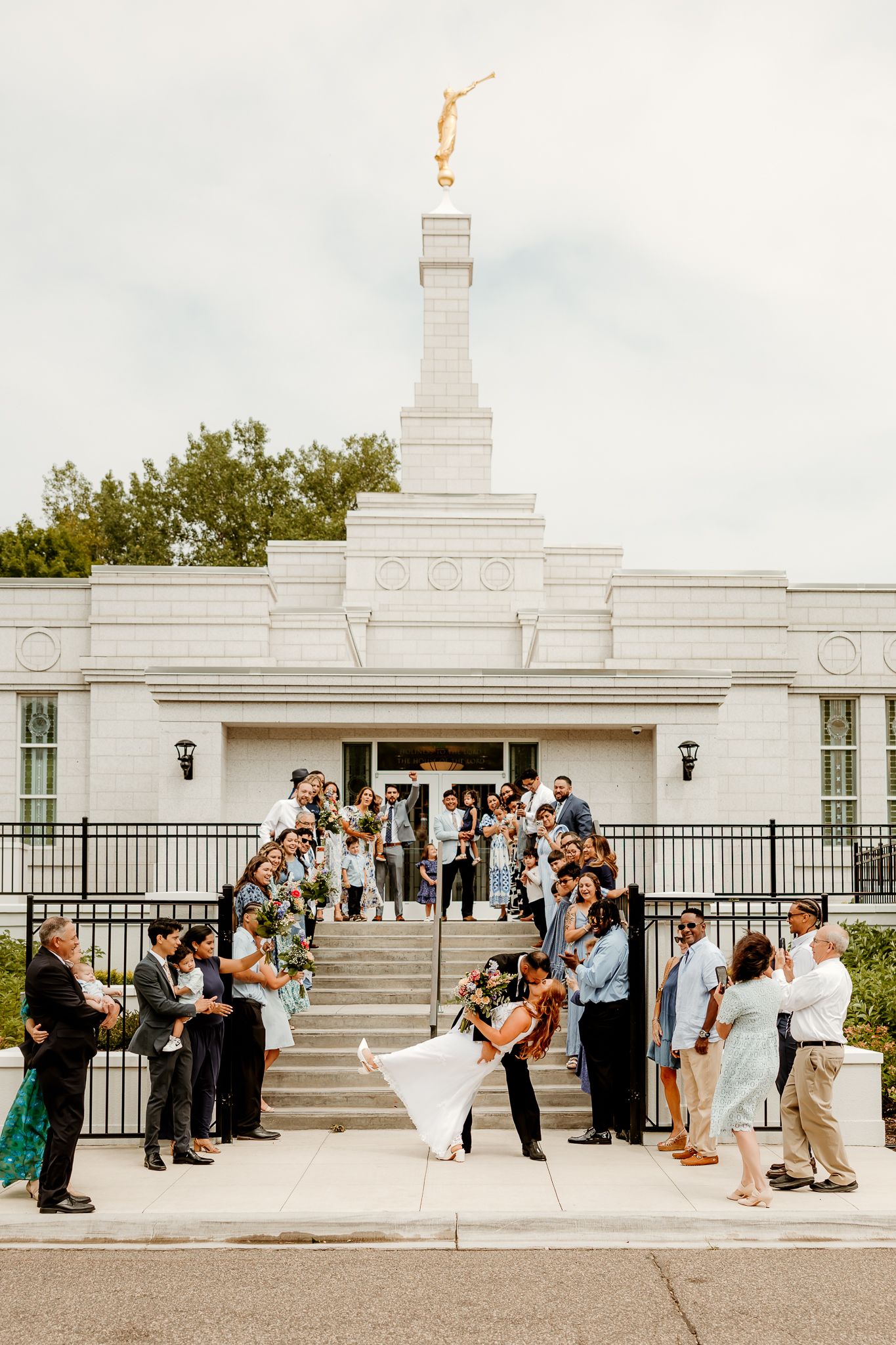 Newlyweds share a dip kiss outside a white LDS temple surrounded by cheering wedding guests on the steps.