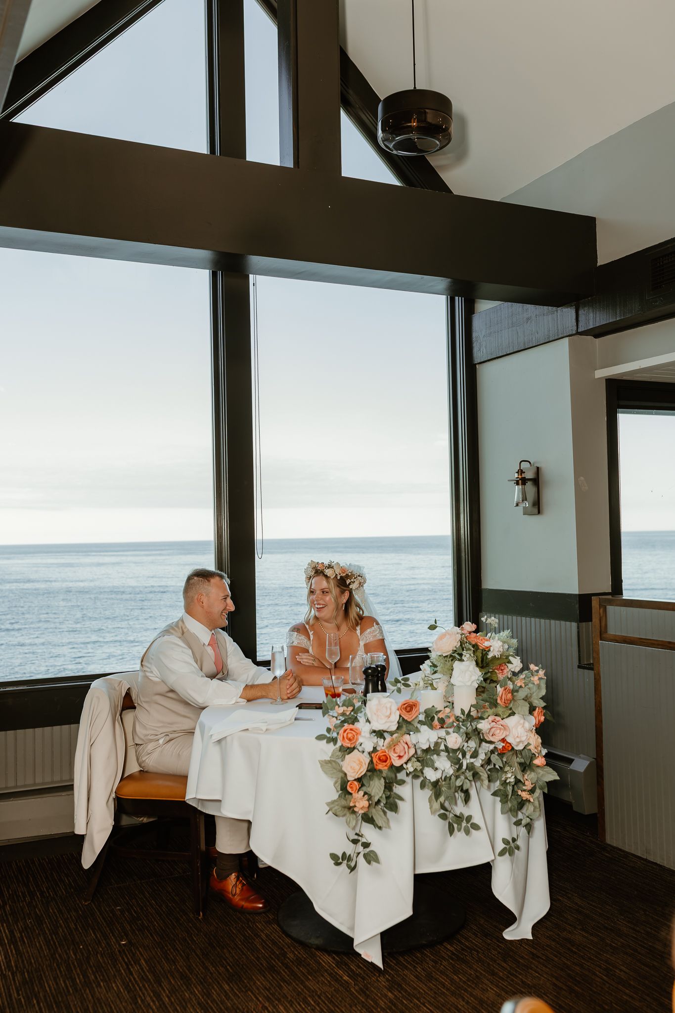 Bride and groom sit at floral-adorned sweetheart table with ocean view backdrop in elegant coastal venue.