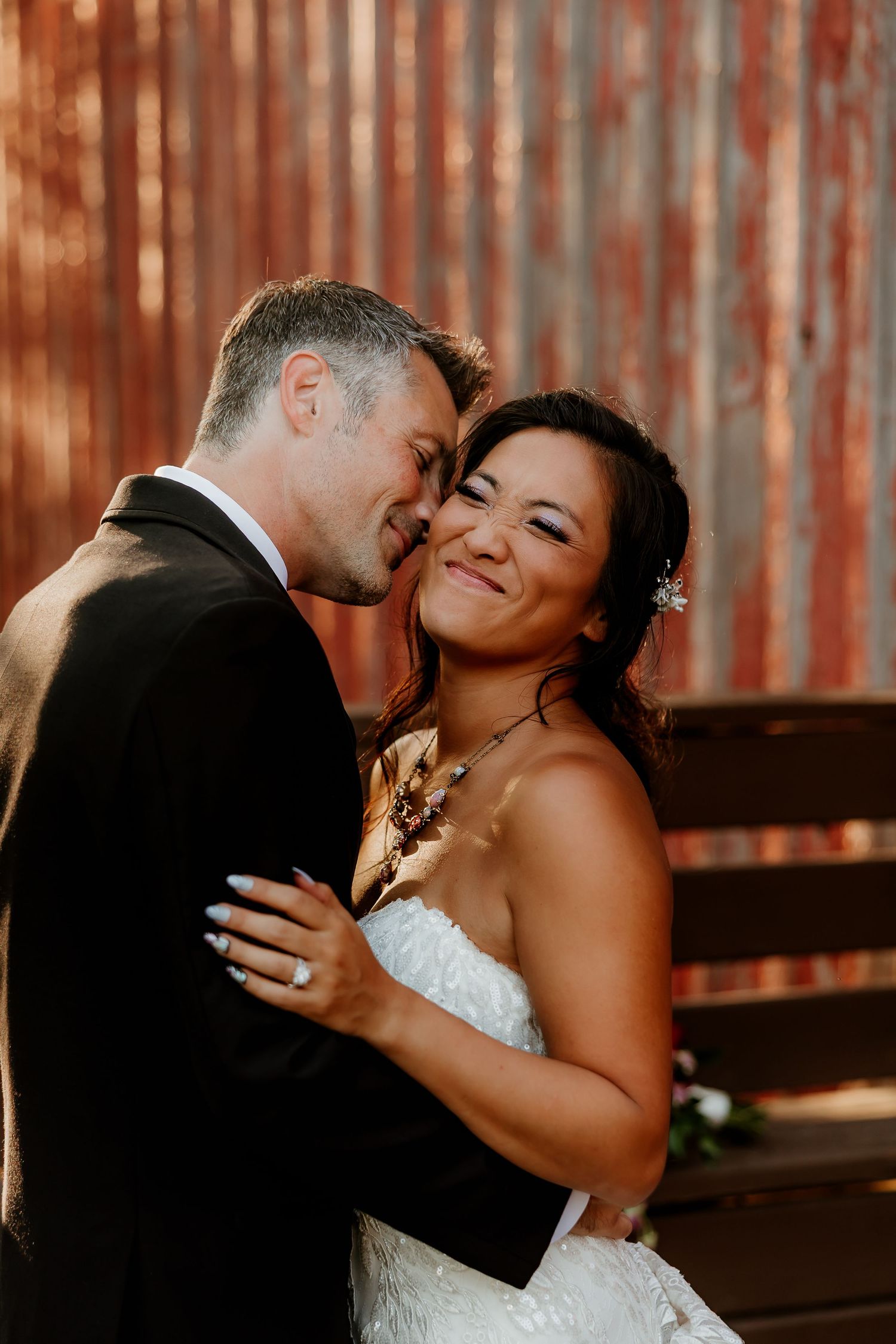 A bride and groom share a romantic embrace in front of a rustic corrugated metal wall on their wedding day.