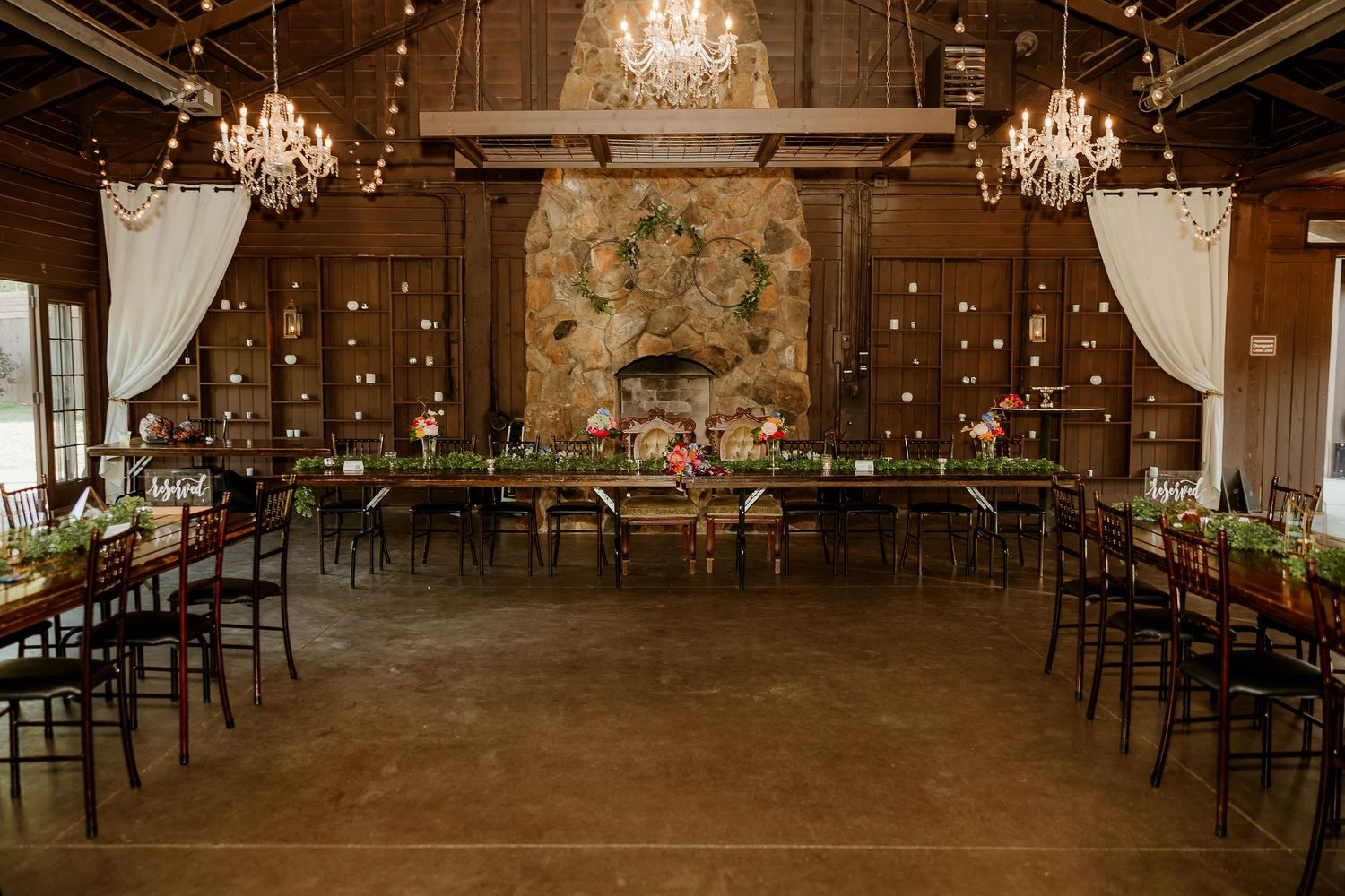 Elegant rustic barn wedding reception with long tables, chiavari chairs, chandeliers, floral centerpieces, and a stone fireplace backdrop.