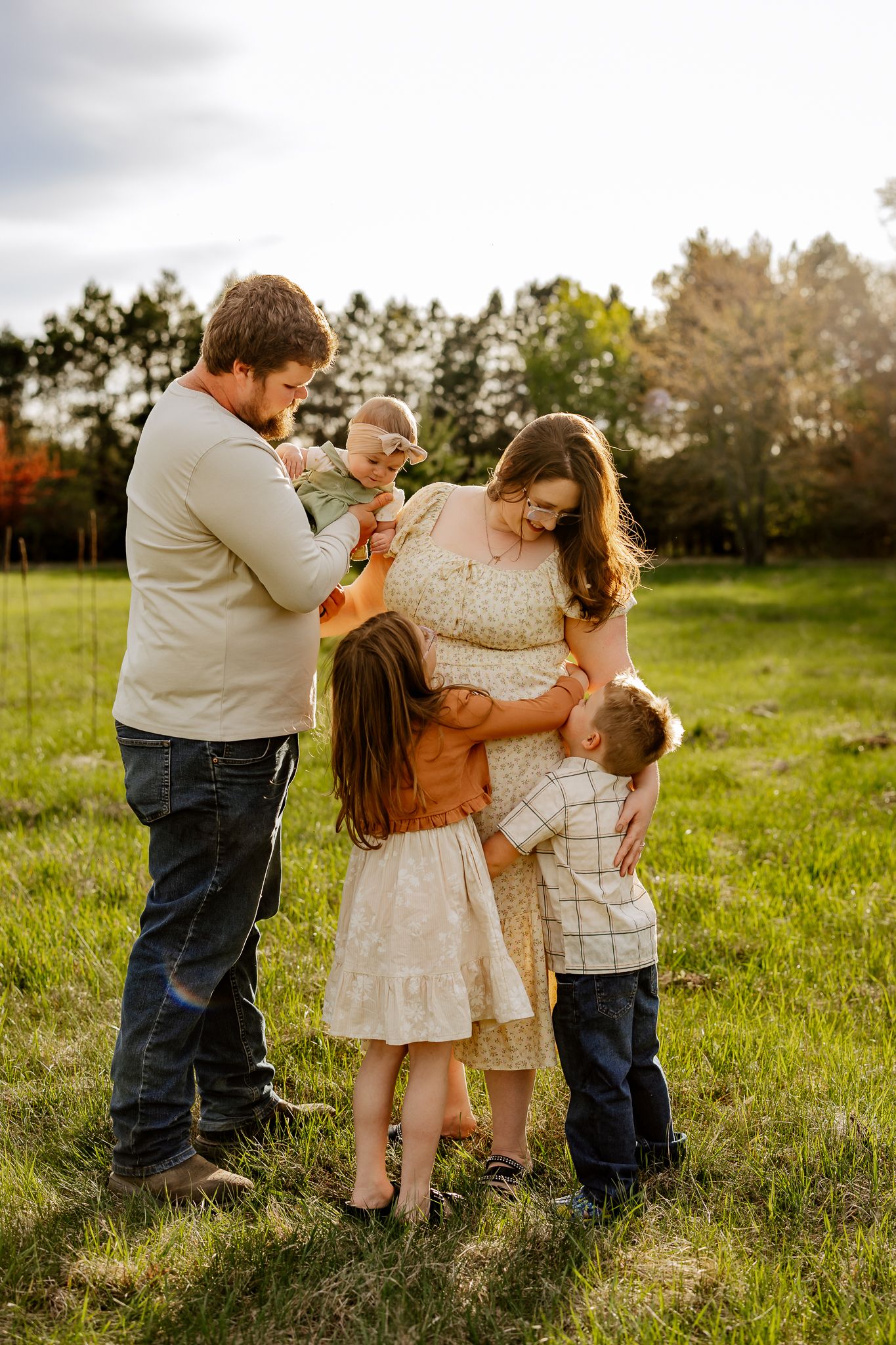 Family of five sharing a warm embrace in a sunlit green field with autumn trees in the background.