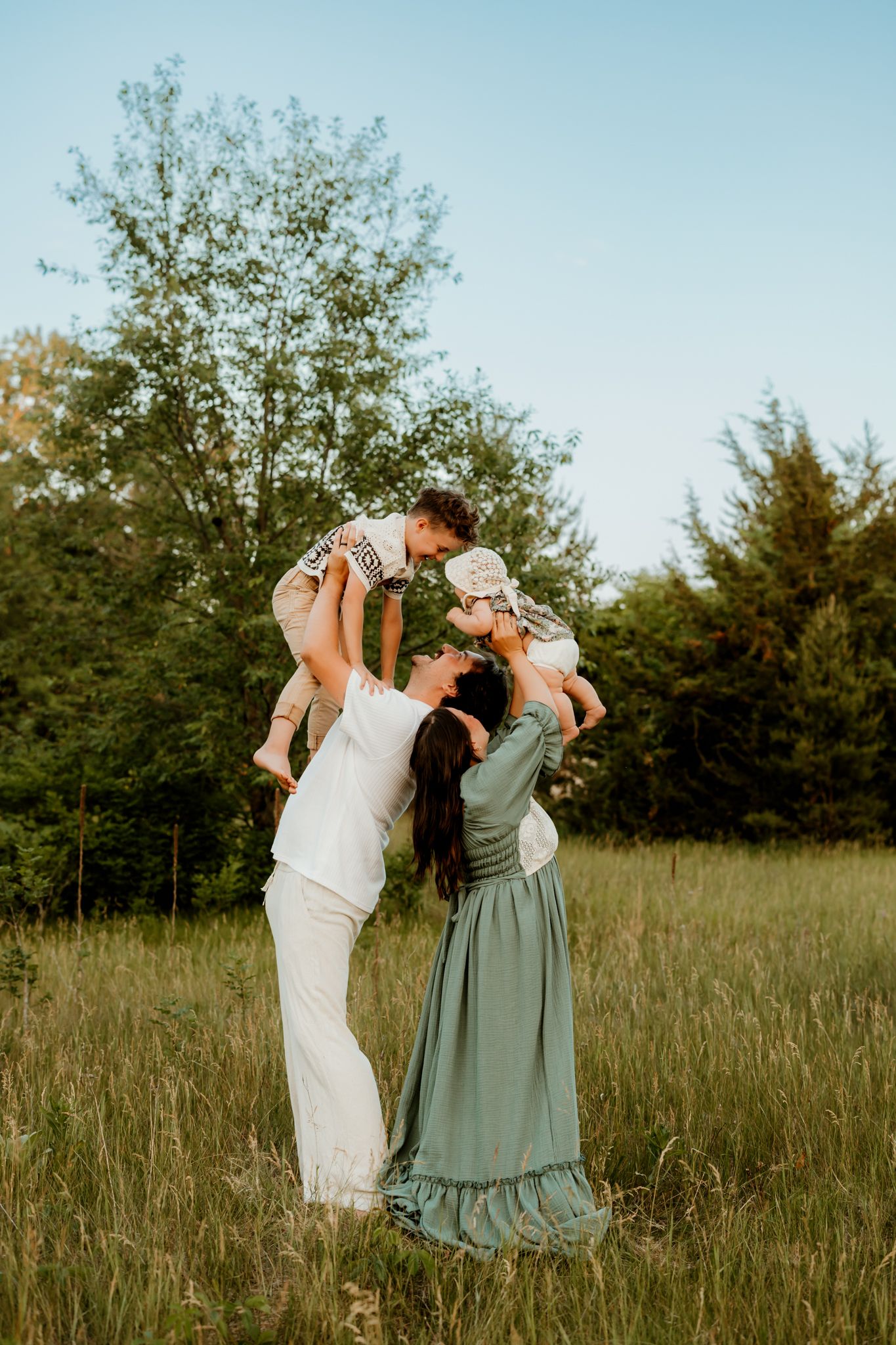 Two women lifting a baby in a sunny green meadow, surrounded by trees, in a joyful outdoor family photo.