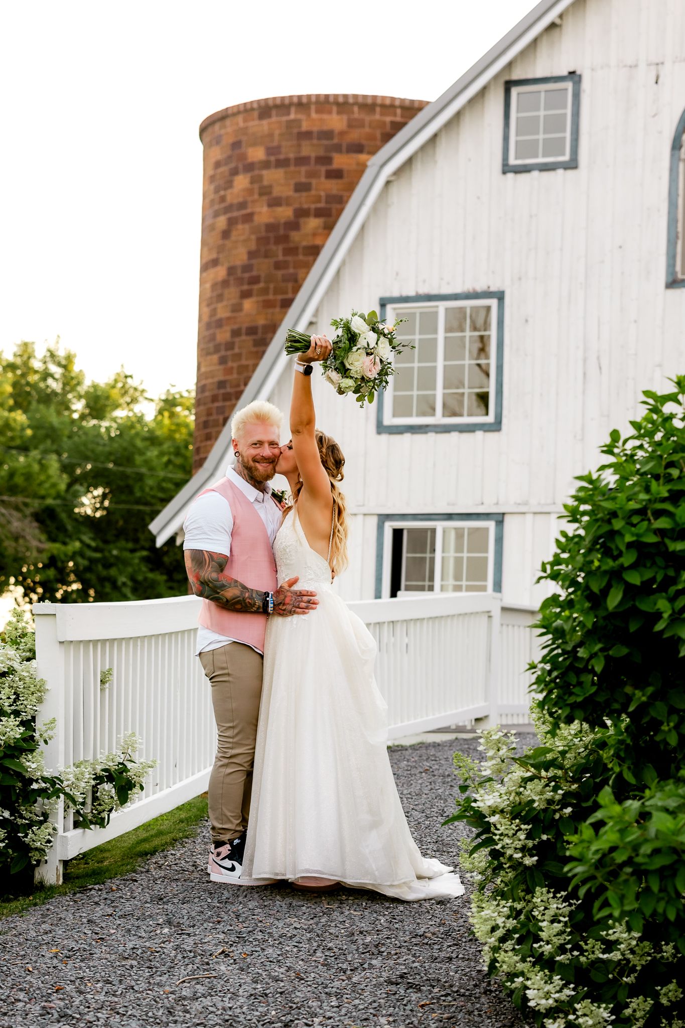 Bride raises bouquet while groom embraces her outside a white barn with silo at a rustic wedding venue.