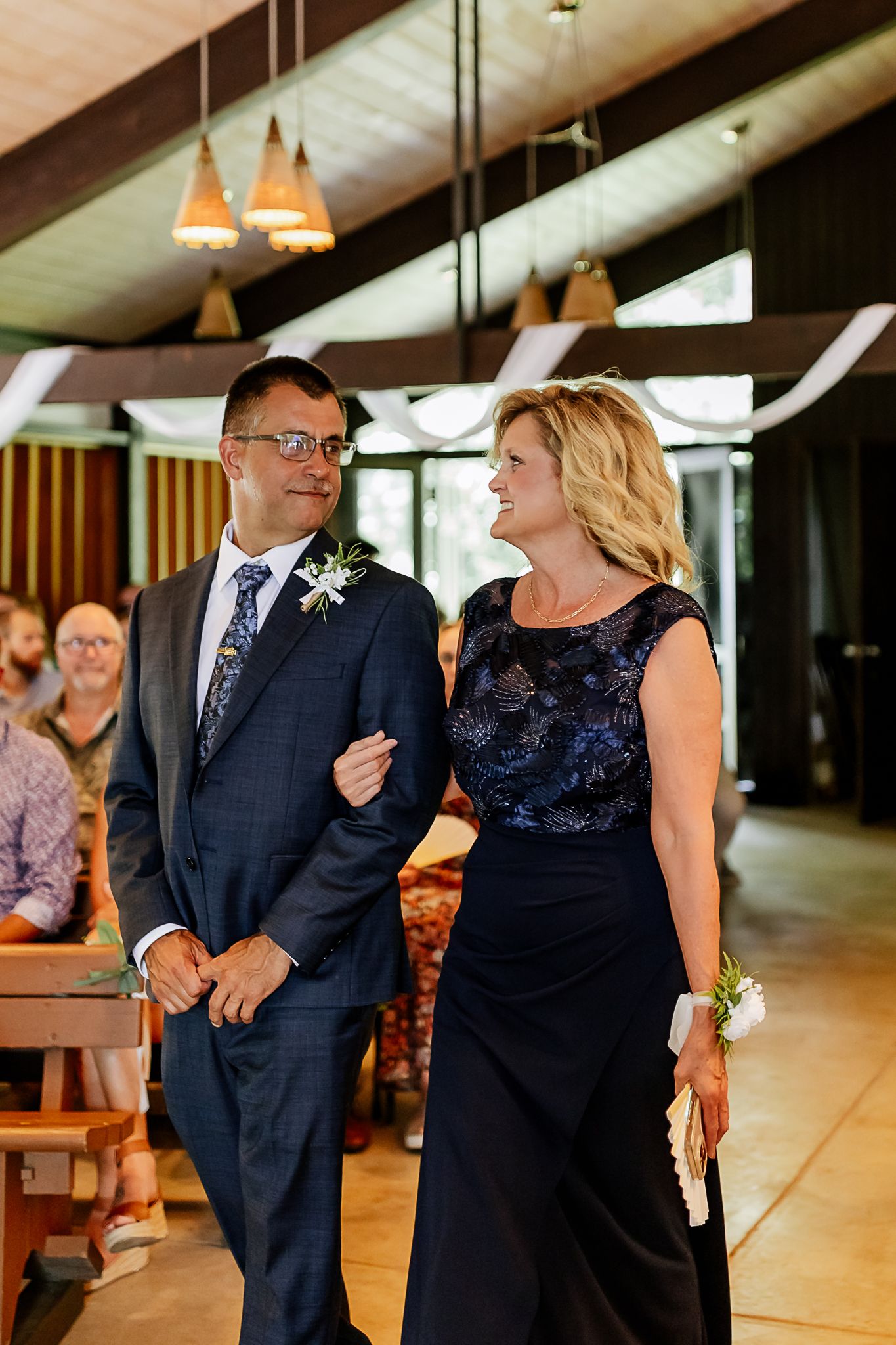A couple walks arm-in-arm down the wedding aisle, man in navy suit with boutonniere, woman in navy lace gown.