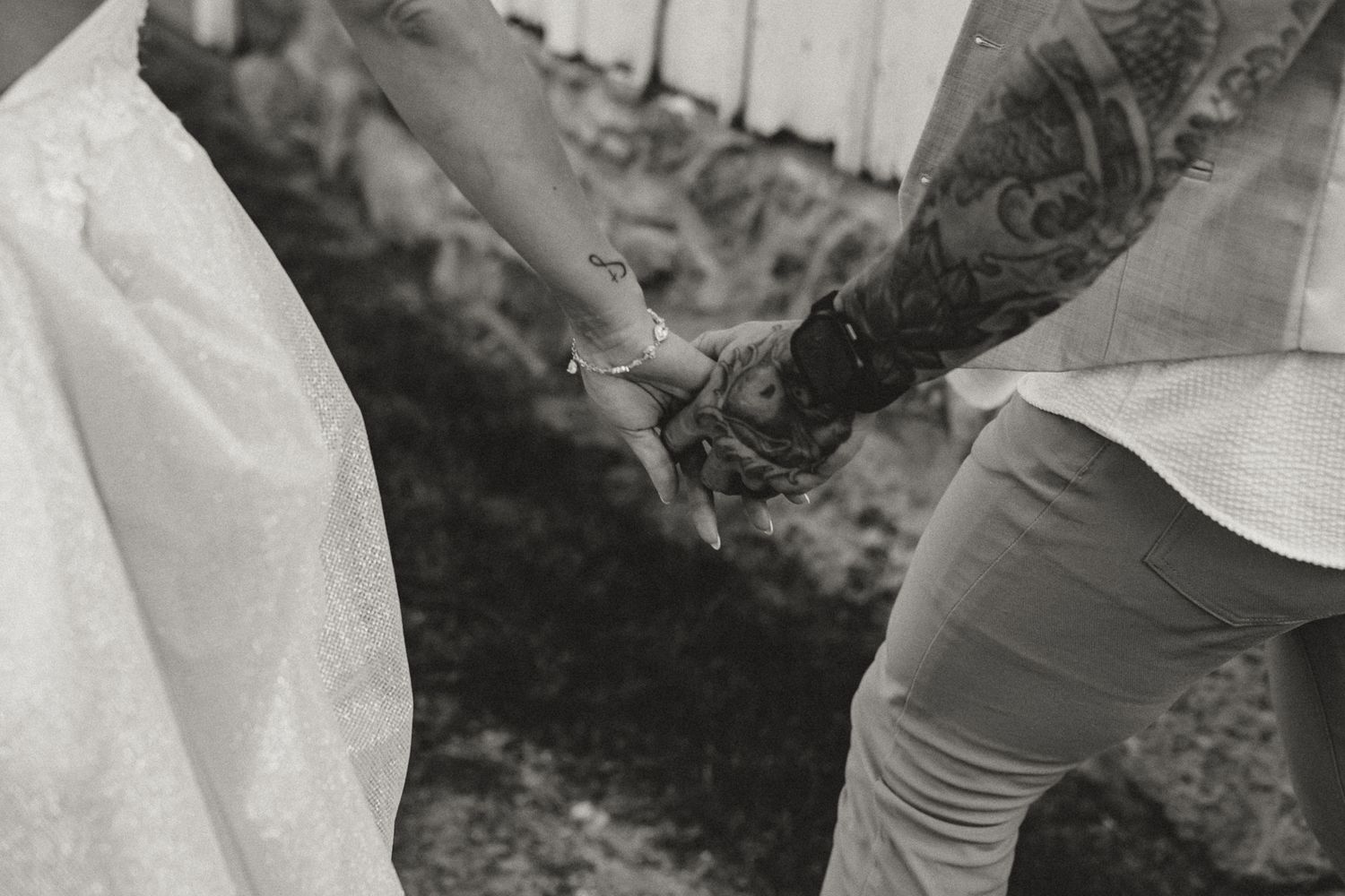 Black and white photo of a tattooed groom holding hands with a bride in a white dress at a wedding.