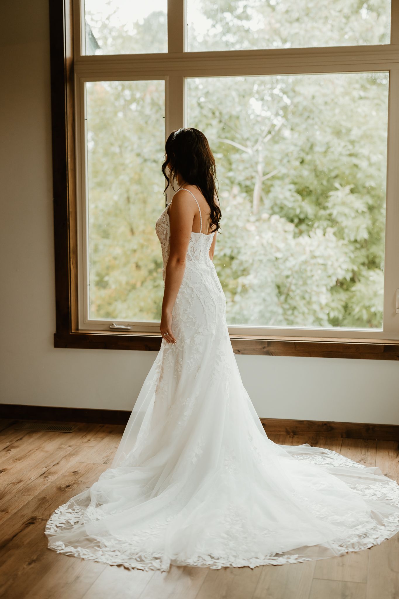 Bride in a lace wedding gown with a long train standing by a large window overlooking green trees.