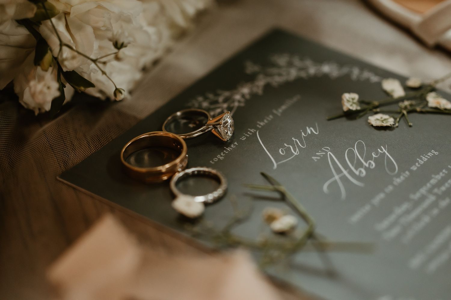 Gold and silver wedding rings with a diamond engagement ring rest on a dark wedding invitation with white florals and greenery.