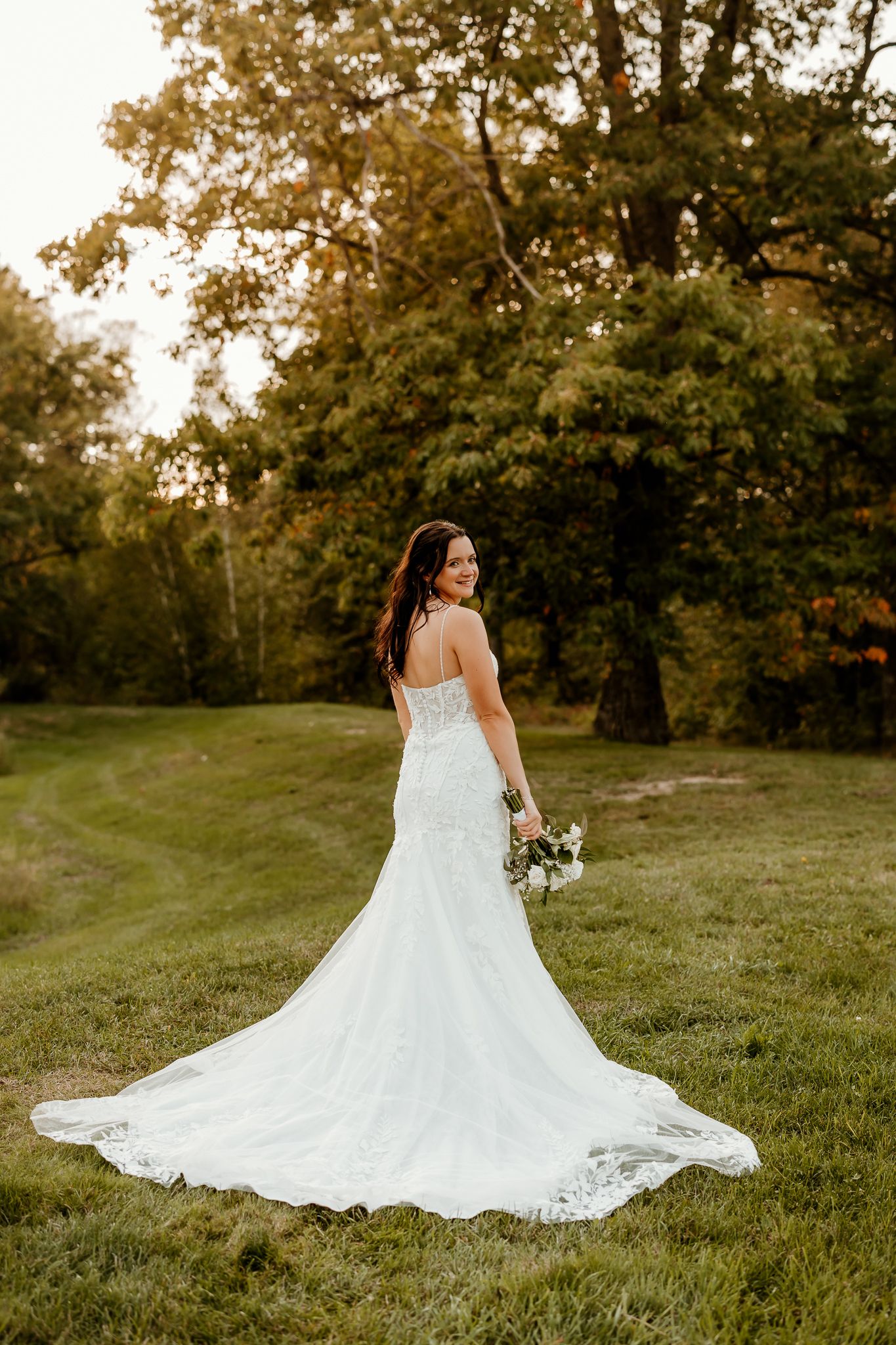 Bride in a white lace mermaid wedding gown with a long train holds a bouquet while smiling over her shoulder on a grassy hill with trees.