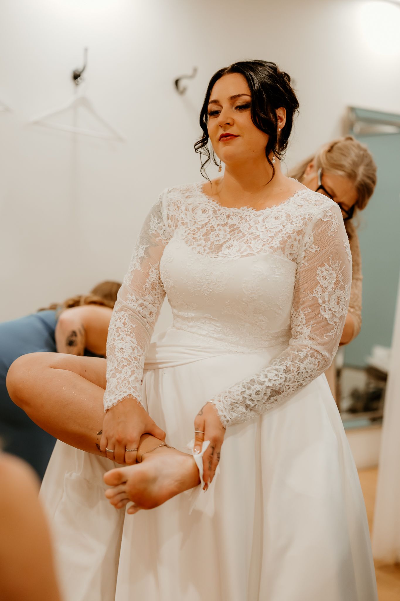 Bride in a white lace long-sleeve wedding dress lifts her foot while bridesmaids help her get ready before the ceremony.