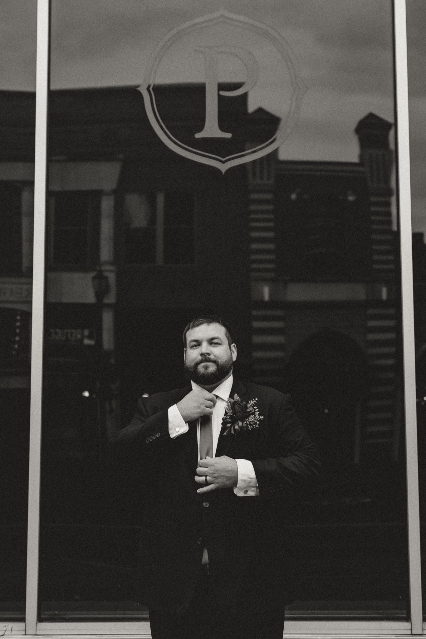 Black and white portrait of a bearded groom in a dark suit with a boutonniere adjusting his tie in front of a glass storefront.
