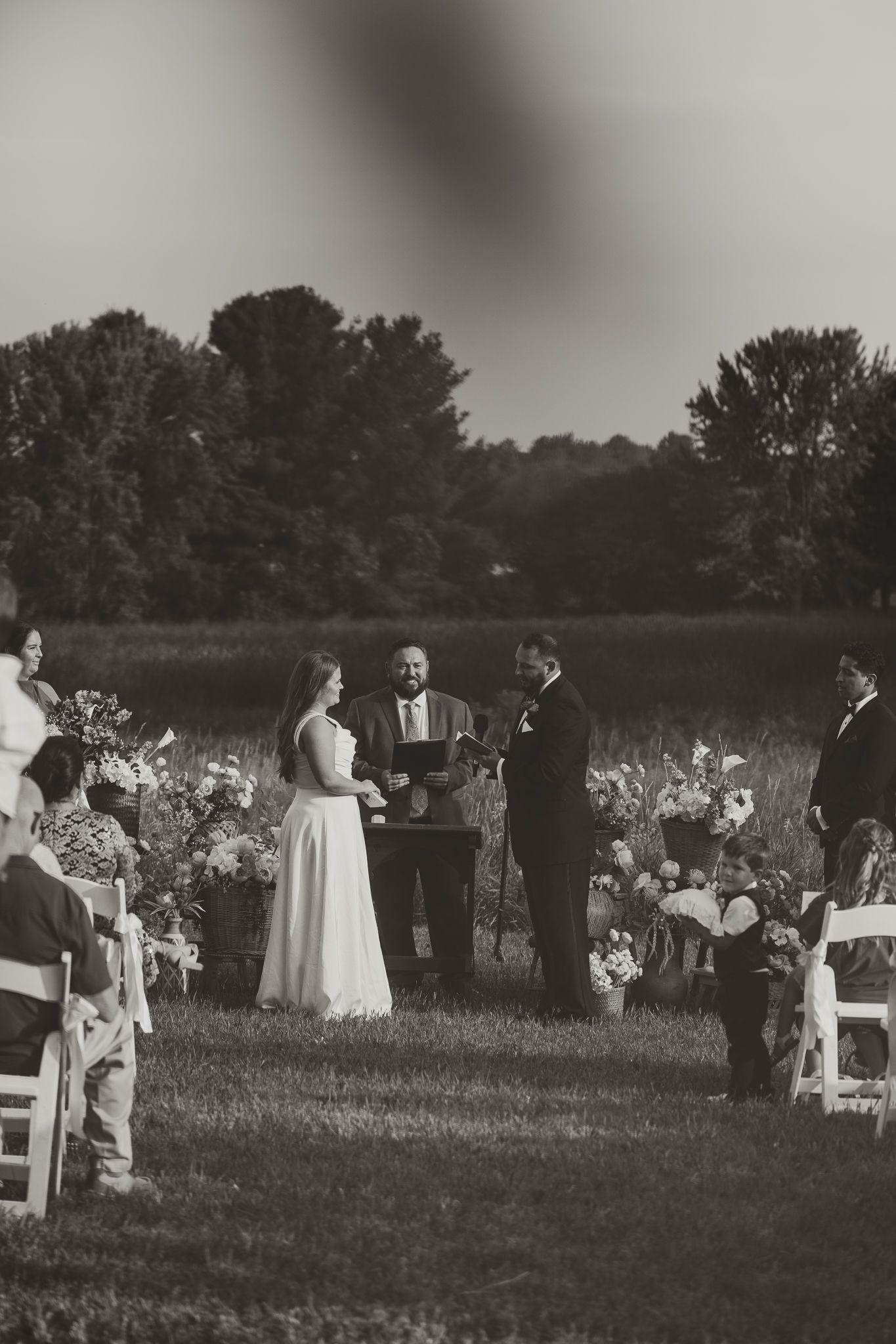 Black and white outdoor wedding ceremony with bride and groom at altar, surrounded by floral arrangements and seated guests.