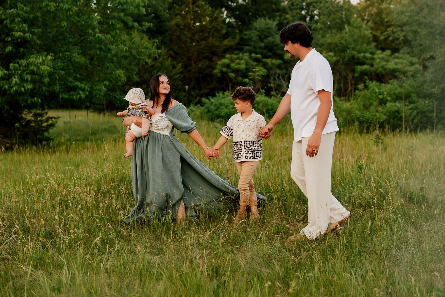 Family of four walking hand in hand through a green meadow at golden hour.