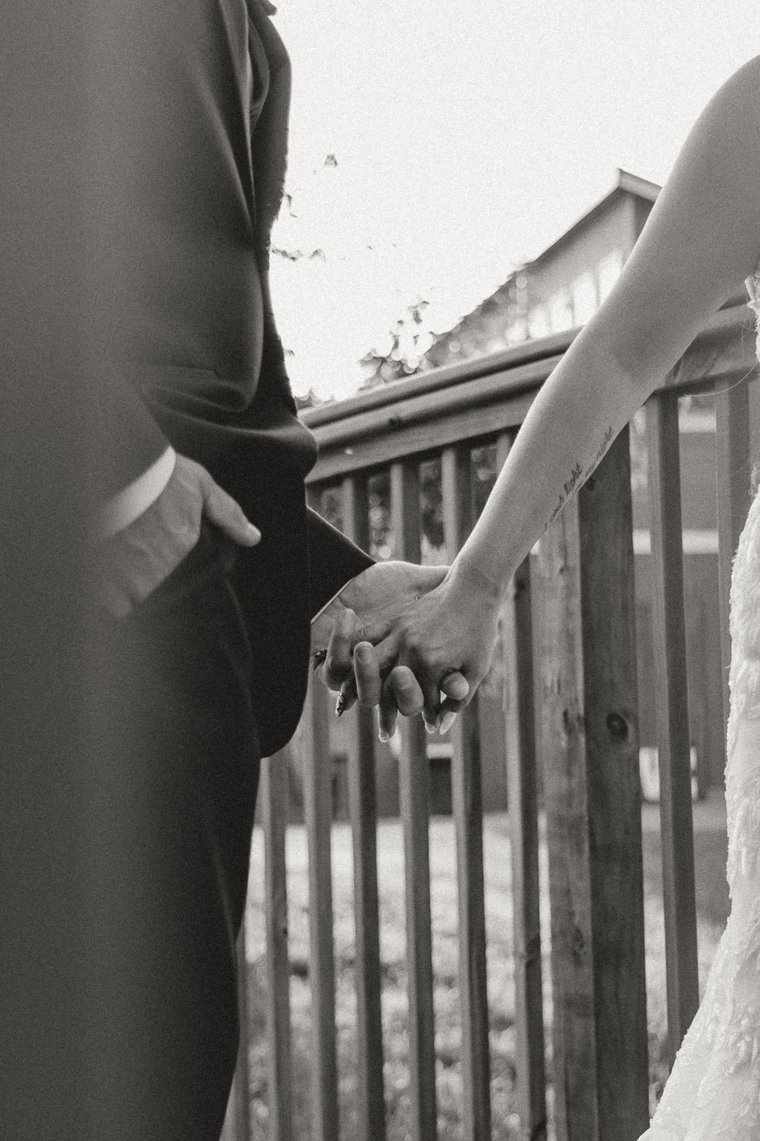 Black and white photo of a bride and groom holding hands at their wedding ceremony near a wooden fence.