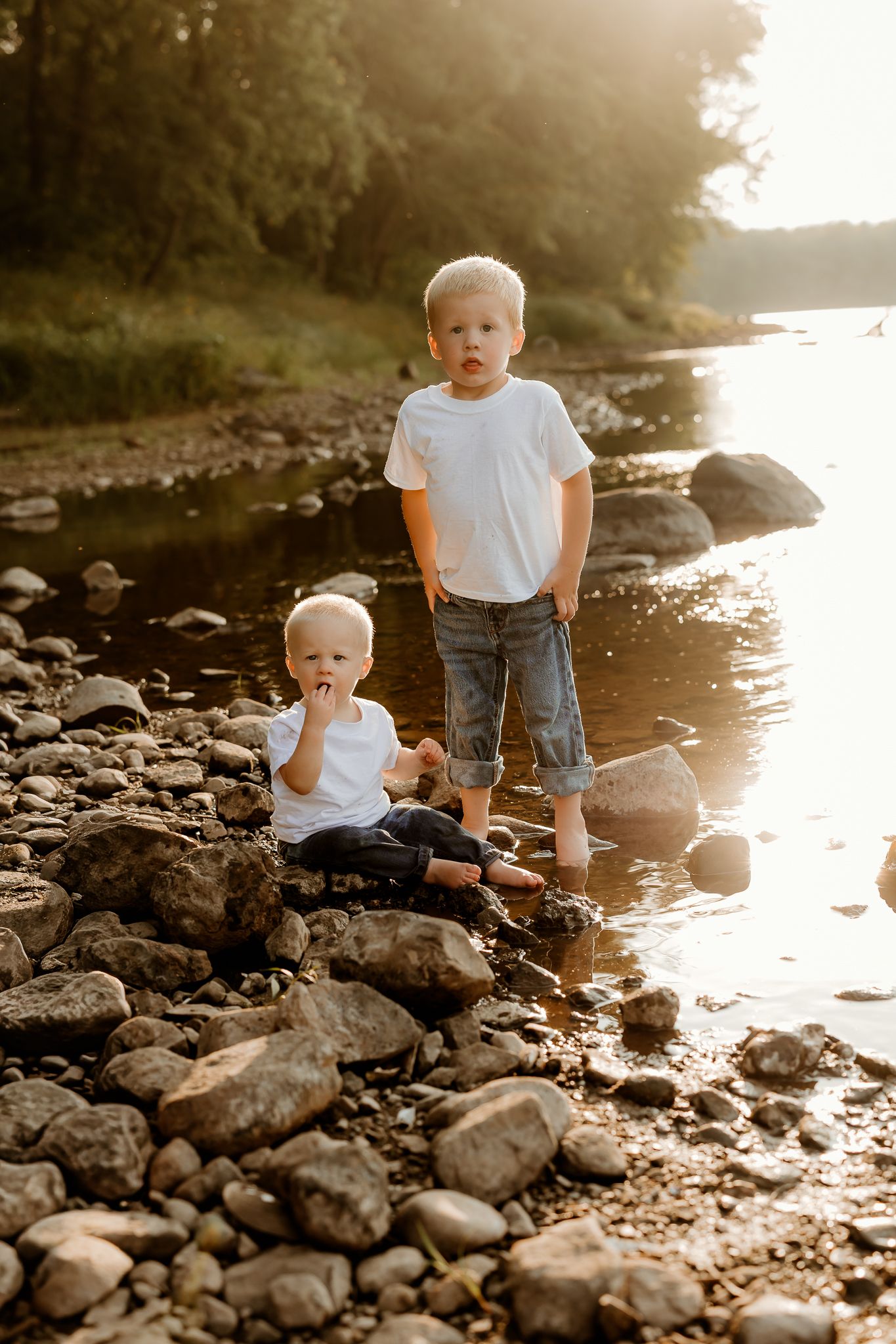 Two young blonde boys in white t-shirts playing on rocky riverbank at golden sunset hour.