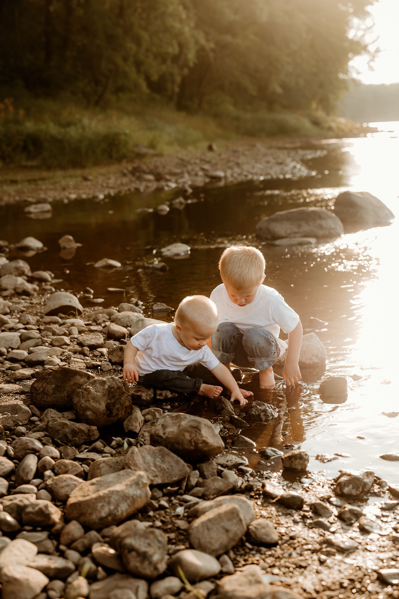 Two young boys in white shirts playing with rocks along a sunlit river shore at golden hour.