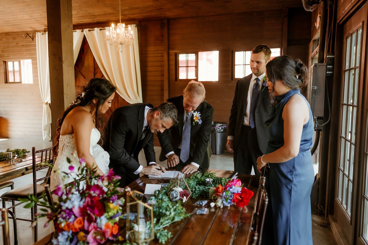 Wedding party signing marriage certificate at rustic wooden table adorned with colorful floral arrangements in a barn venue.