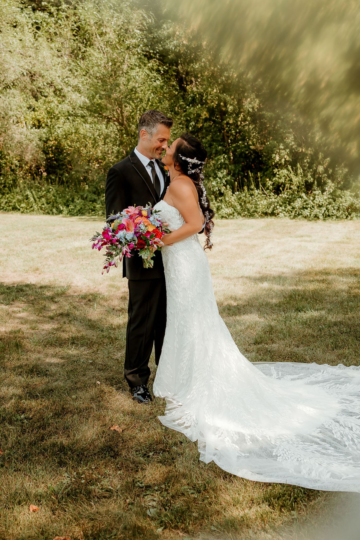 Bride in lace gown with colorful bouquet and groom in black tuxedo sharing a romantic moment outdoors.