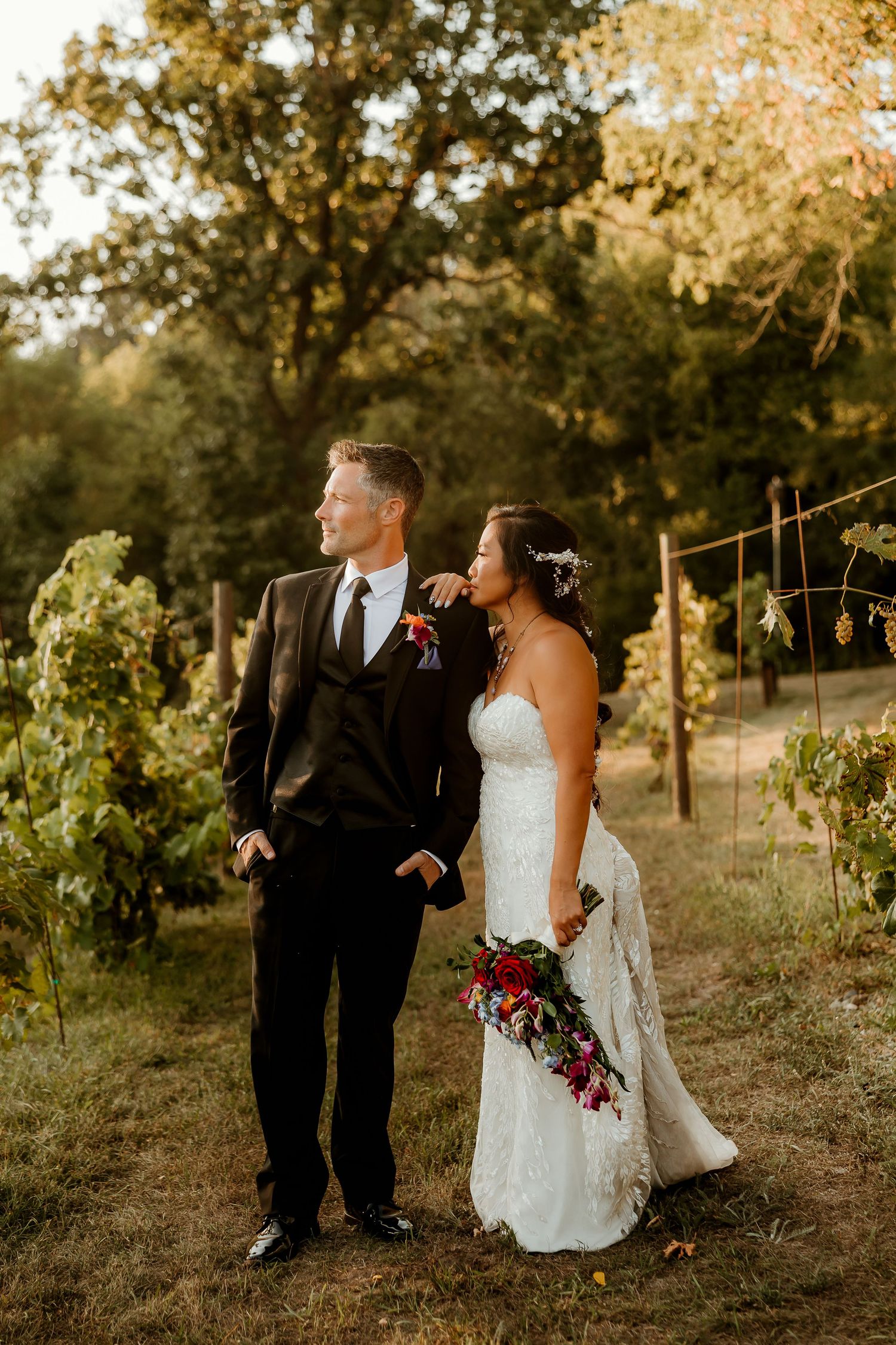 Bride and groom stand together in a vineyard at golden hour, she holds a red bouquet in a lace gown, he wears a black suit.