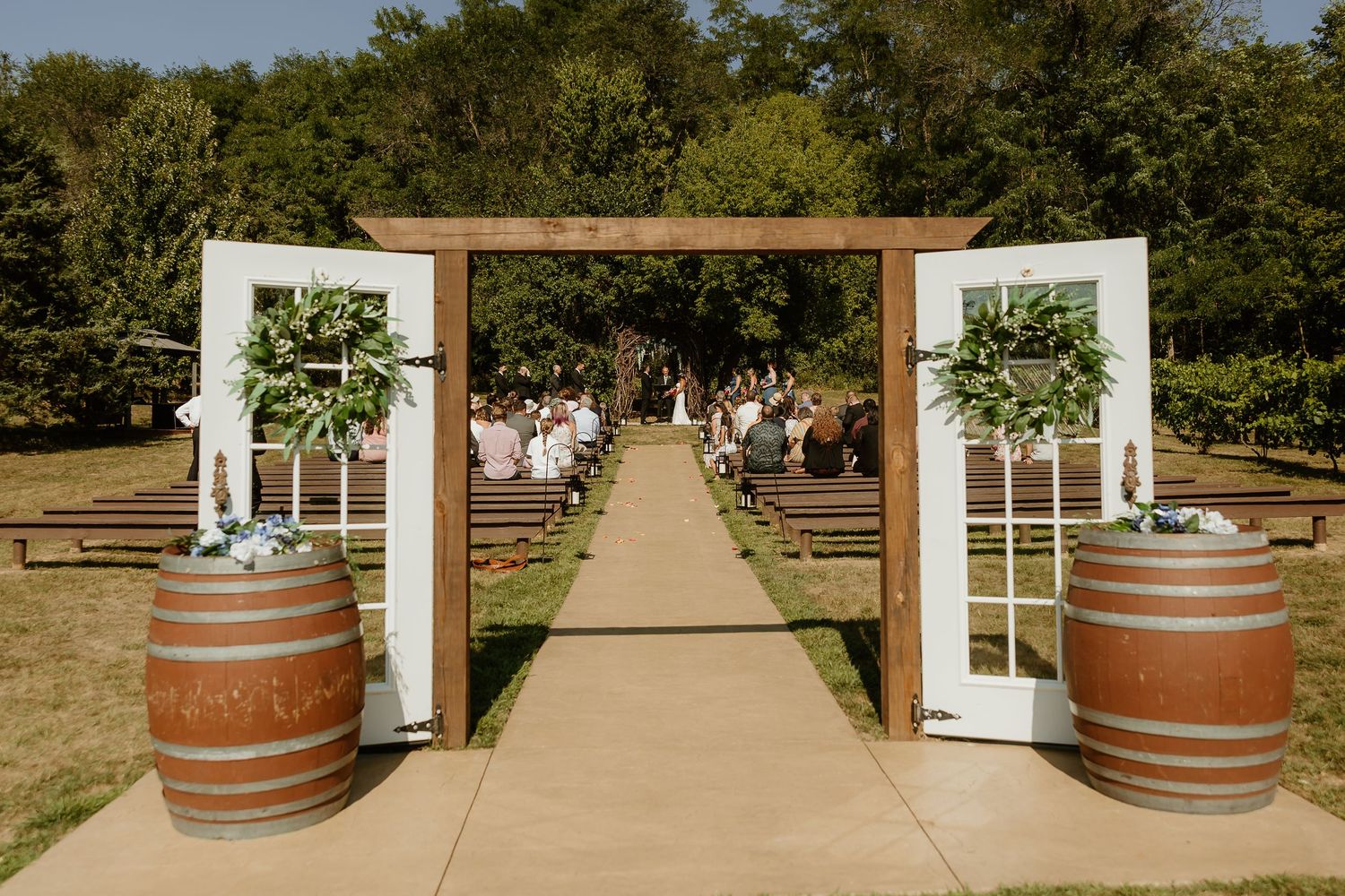 Rustic outdoor wedding ceremony aisle with wooden barrel decor, white doors, floral arch, and guests seated on benches.