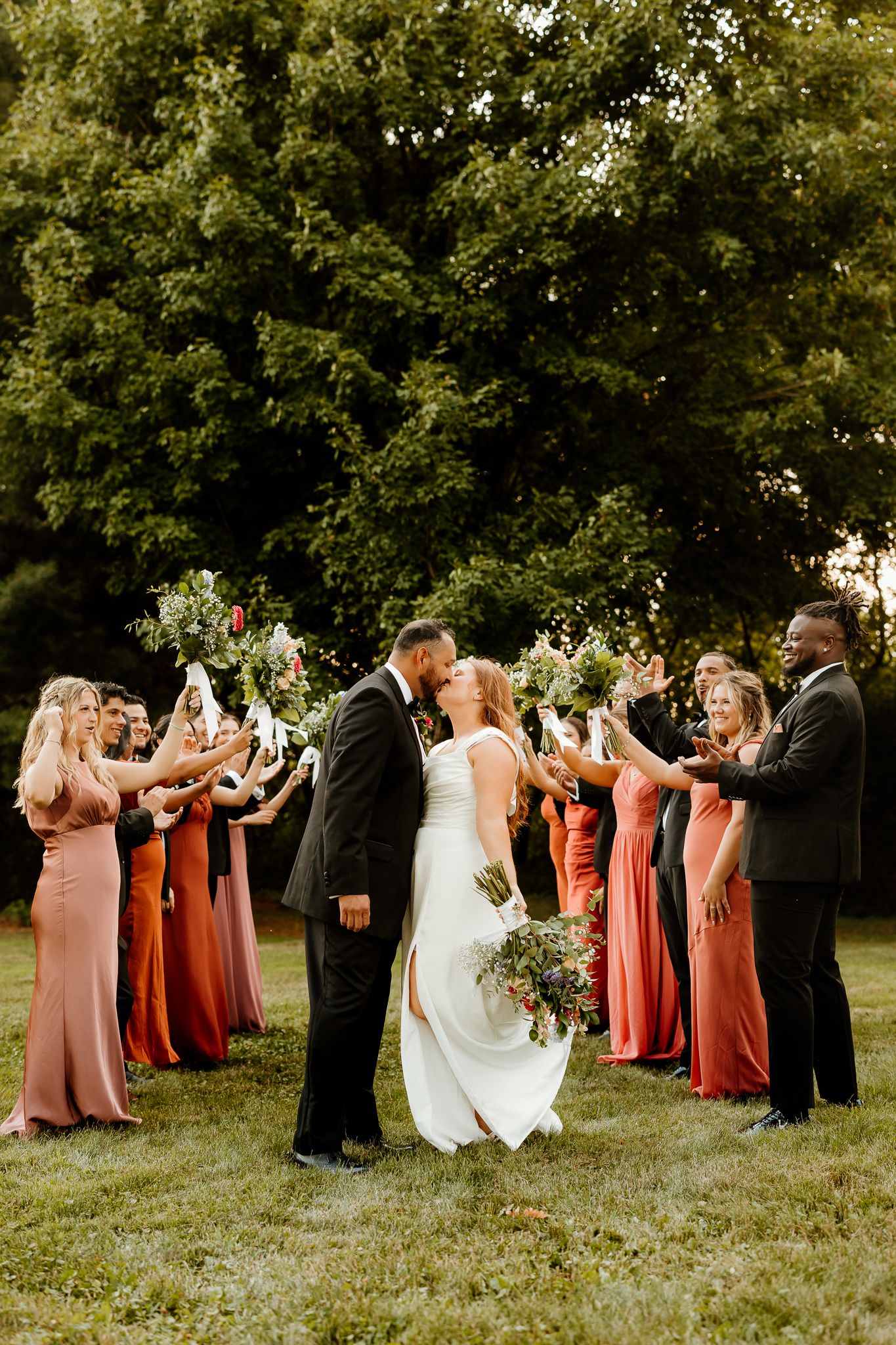 Bride and groom share a kiss while wedding party in rust and mauve gowns raises bouquets in celebration outdoors.