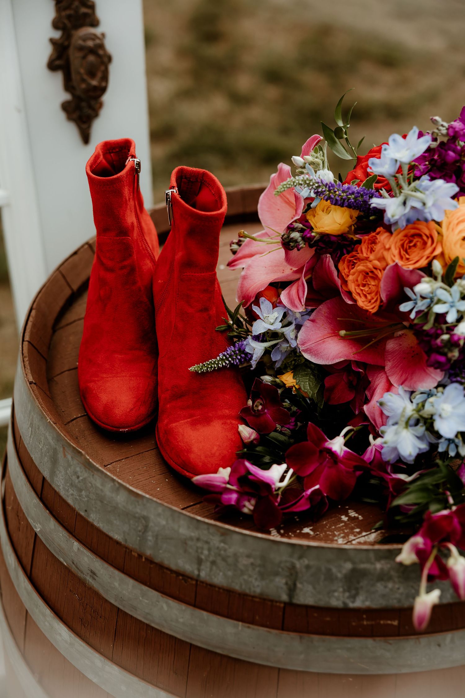 Red ankle boots and colorful bridal bouquet with roses and orchids displayed on a rustic wooden barrel.