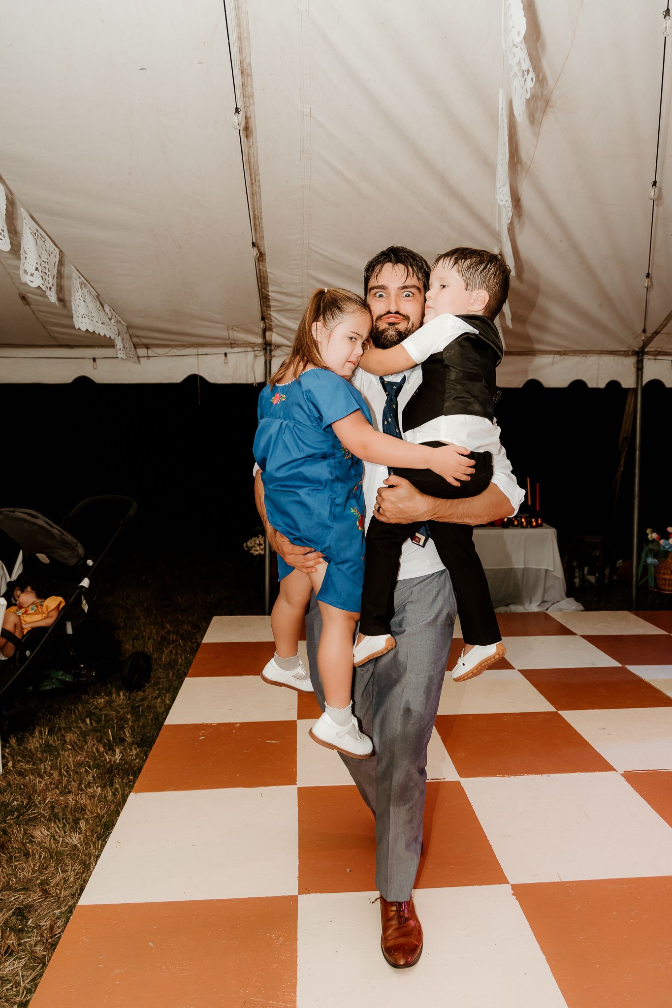 Man in suit holding two children on a checkered dance floor inside a wedding reception tent.