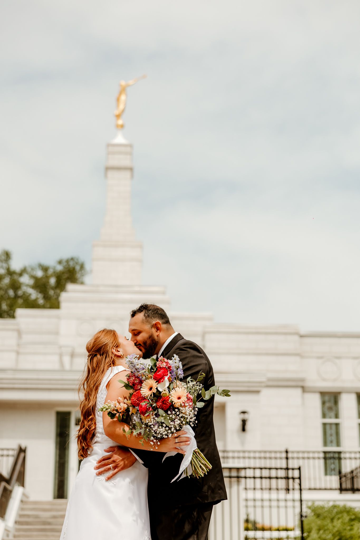 Bride and groom share a romantic kiss outside a white LDS temple, bride holding a colorful bouquet.