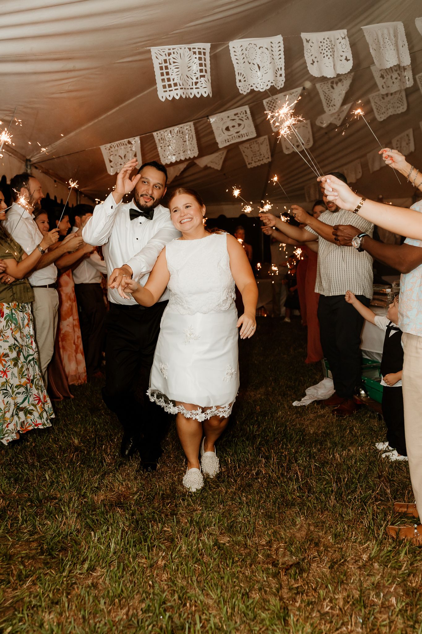 Bride and groom walk through sparkler send-off under a tent decorated with white papel picado at their outdoor wedding reception.