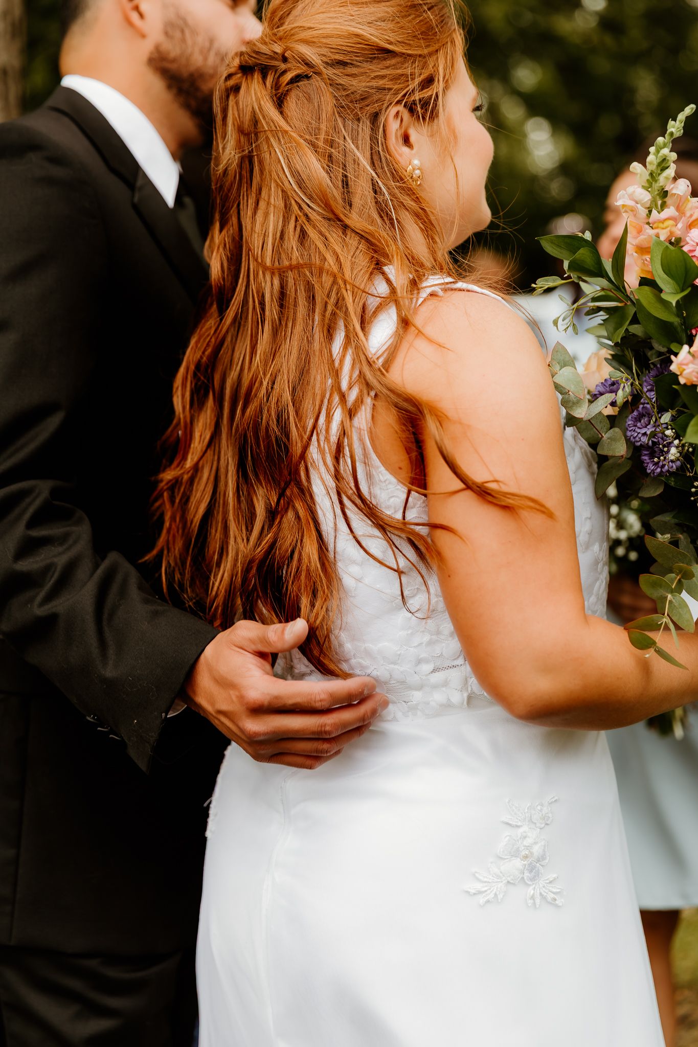 Bride in white dress with long red hair holds bouquet while groom in black suit embraces her waist outdoors.