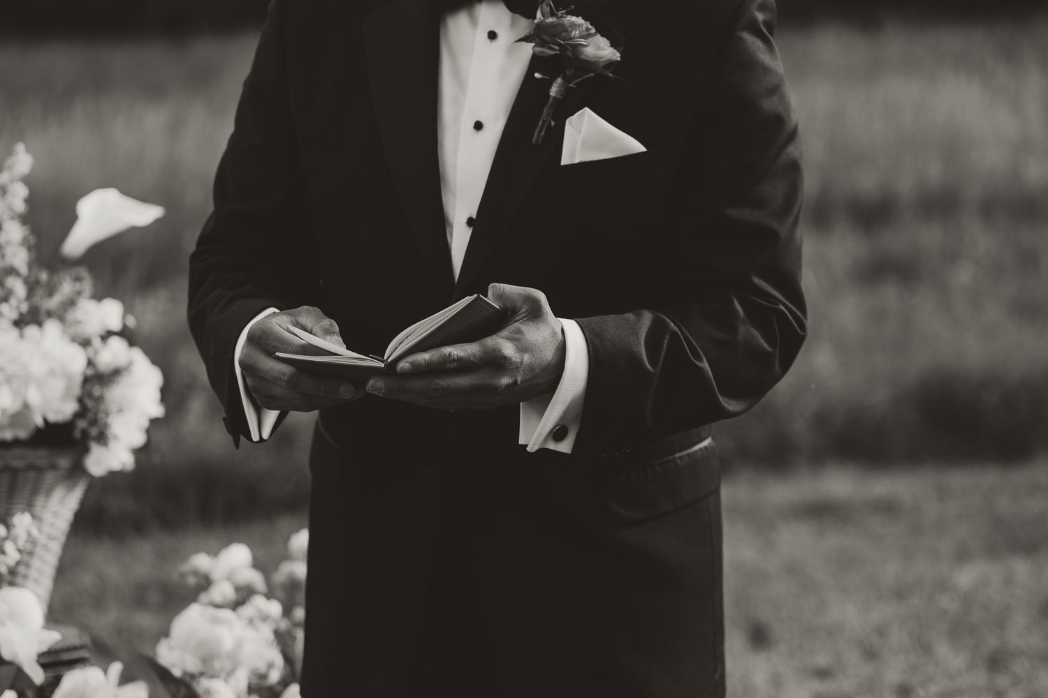Groom in tuxedo reading vows from a book during outdoor wedding ceremony, black and white photo.