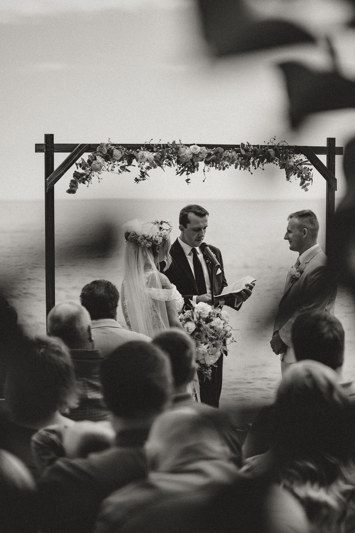Black and white outdoor wedding ceremony with bride and groom at floral arch by the ocean.