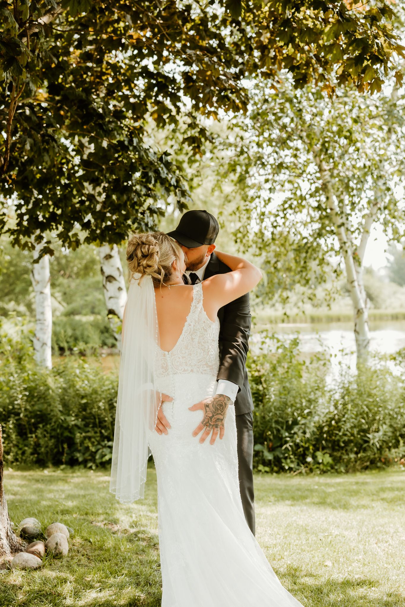 A bride and groom share a romantic kiss outdoors under lush green trees on their wedding day.