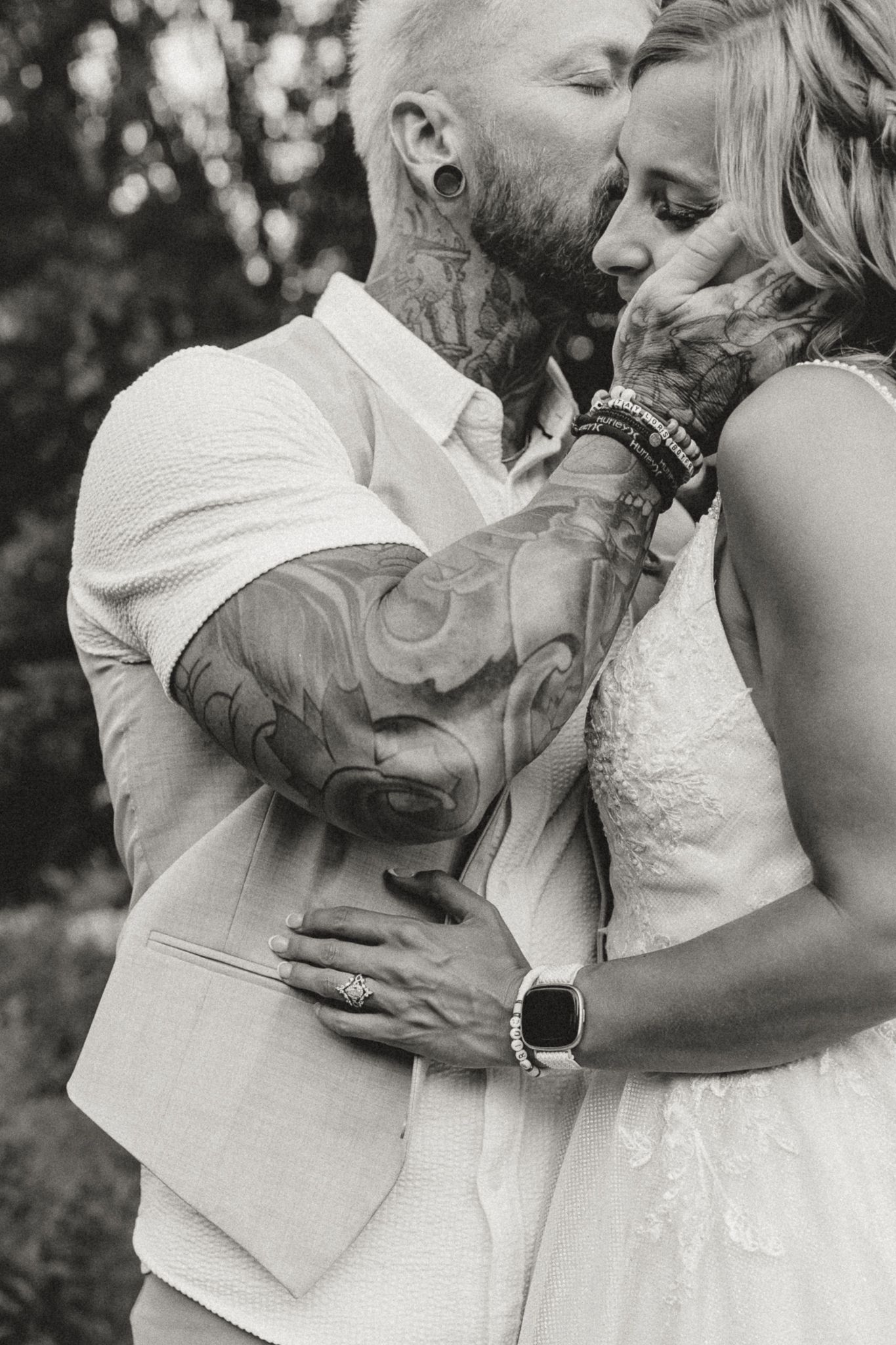 Black and white photo of tattooed groom in white suit tenderly holding bride's face during romantic wedding moment.