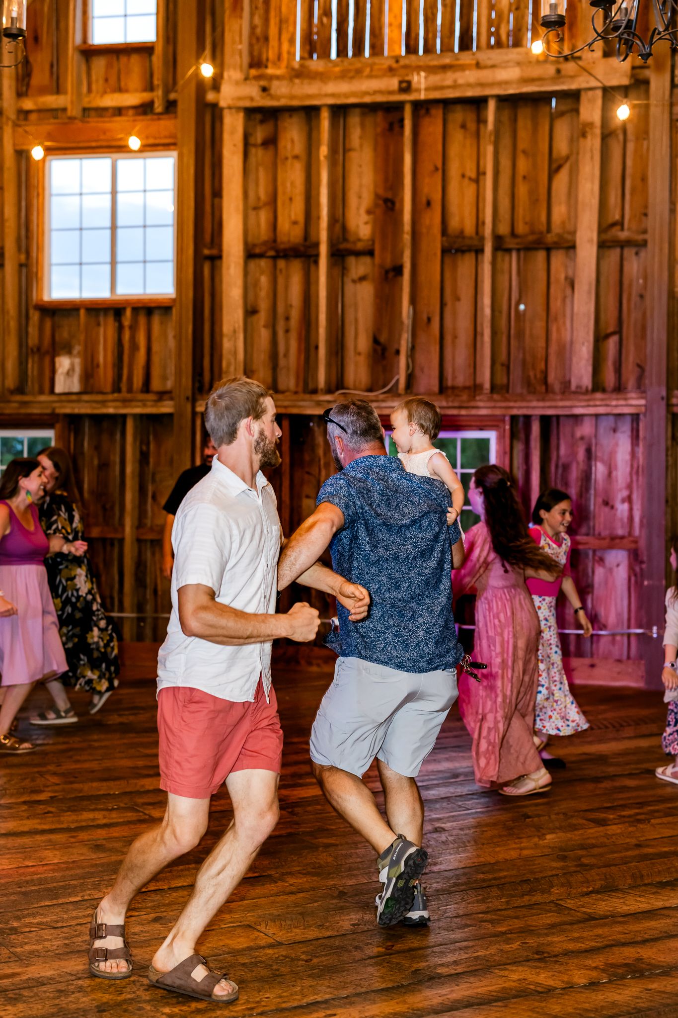 Guests dancing joyfully in a rustic wooden barn venue with string lights during a wedding reception.