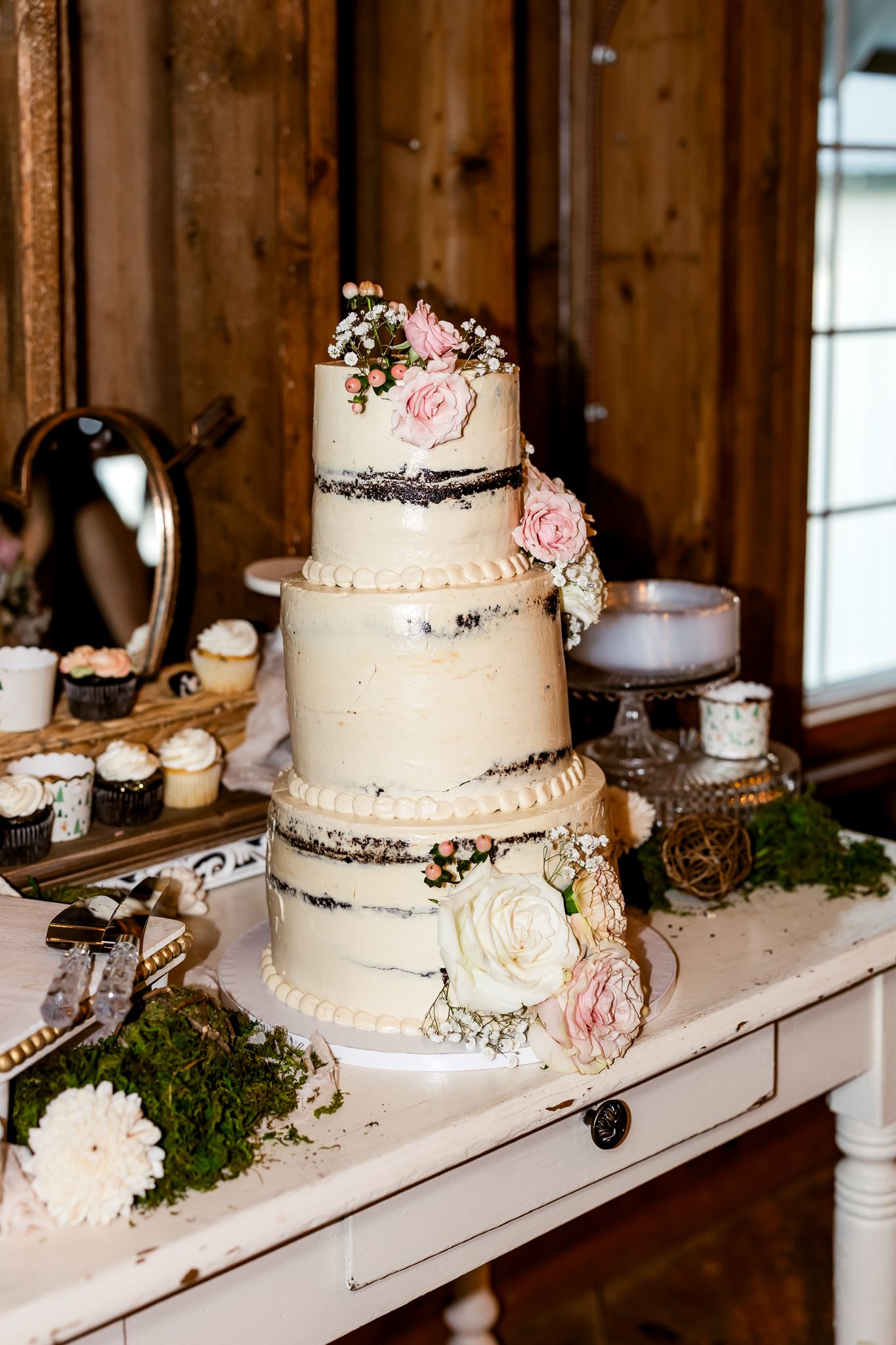 Three-tier semi-naked wedding cake adorned with pink and white roses on a rustic white table.