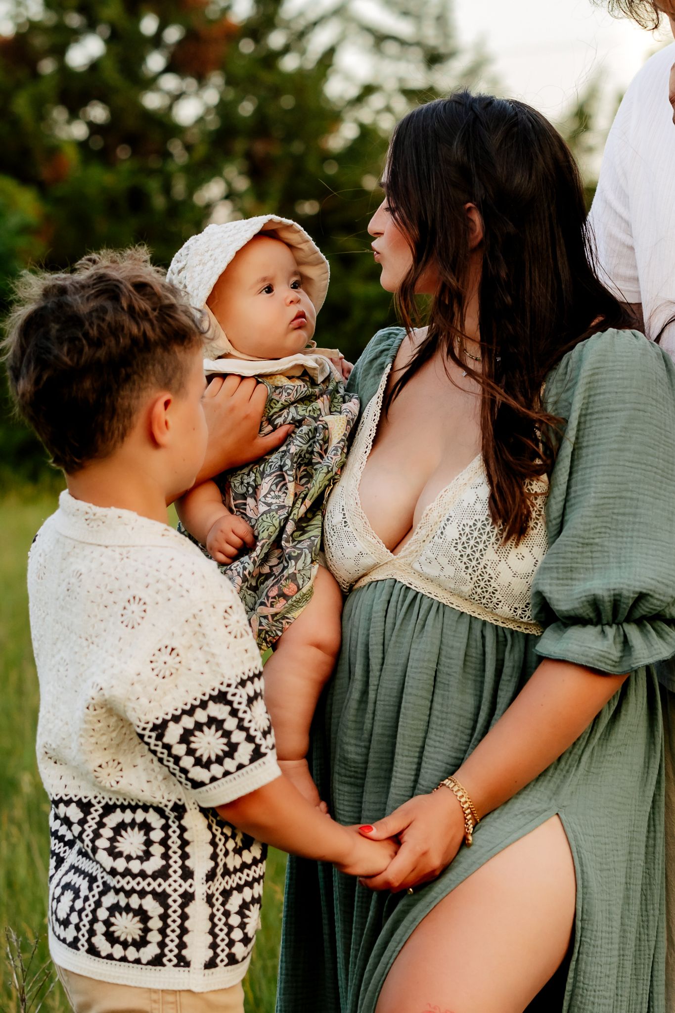 Pregnant mother in sage green dress smiling at baby held by older sibling during outdoor maternity session.